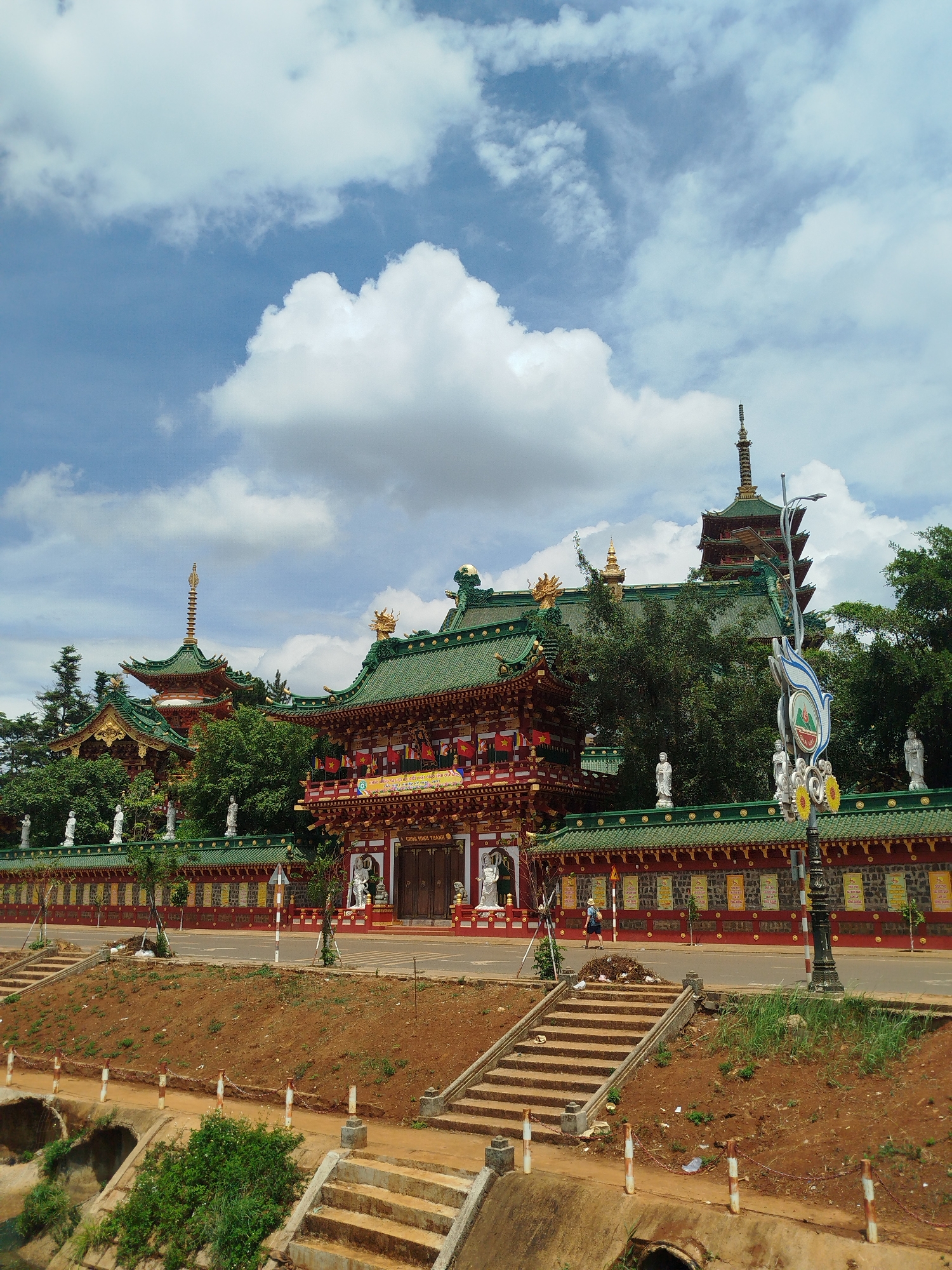 The image depicts a large Buddhist temple complex with multiple structures in Pleiku Ward, Vietnam. The buildings have green roofs, red walls, and intricate golden detailing. Statues line the tops of walls and are placed at building entrances. One individual is walking on the street in front of the temple. A set of concrete steps leads up from a lower level separated by a road. The sky is blue with large, puffy white clouds. The overall environment is bright and sunny. A decorative light post stands to the right of the temple wall.