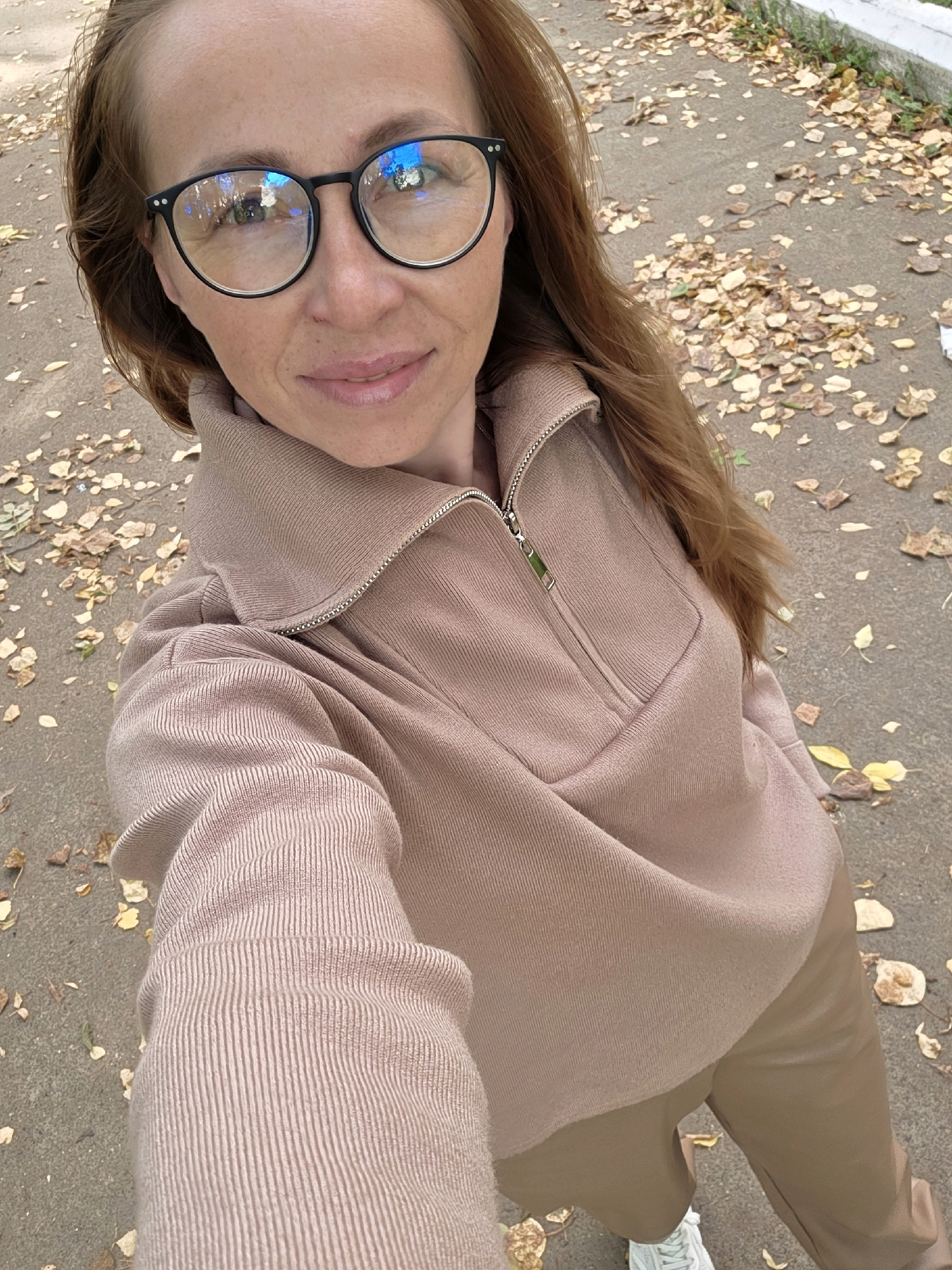A woman with long, auburn hair and wearing glasses is taking a selfie outdoors. She is dressed in a beige ribbed sweatshirt with a quarter-zip collar and beige pants. The background appears to be a paved walkway or road covered with fallen autumn leaves. The environment suggests a park or a residential street in Chaikovsky, Russia, during the fall season.