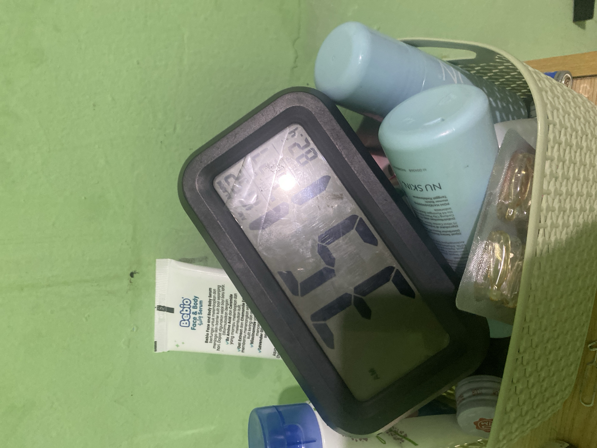 A cluttered shelf or basket contains various items, including skincare products and a digital clock. The clock displays "45" in large digits and "82.9" in smaller digits above, possibly indicating temperature. Among the skincare products are bottles of "NU SKIN", and a tube of "Bebio Face & Body Baby Serum" containing instructions in Indonesian. The "Bebio" tube references "Bx Amino Acid dan Ceramide", "Oat Extract", "Chamomile Extract", "Niacinamide", and "Lavender Oil". The POM NA number indicates that the "NU SKIN" products are produced in the Gedung City Tower Building, 75 West Condes. This product is manufactured in Indonesia, specifically the city of Balikpapan.
