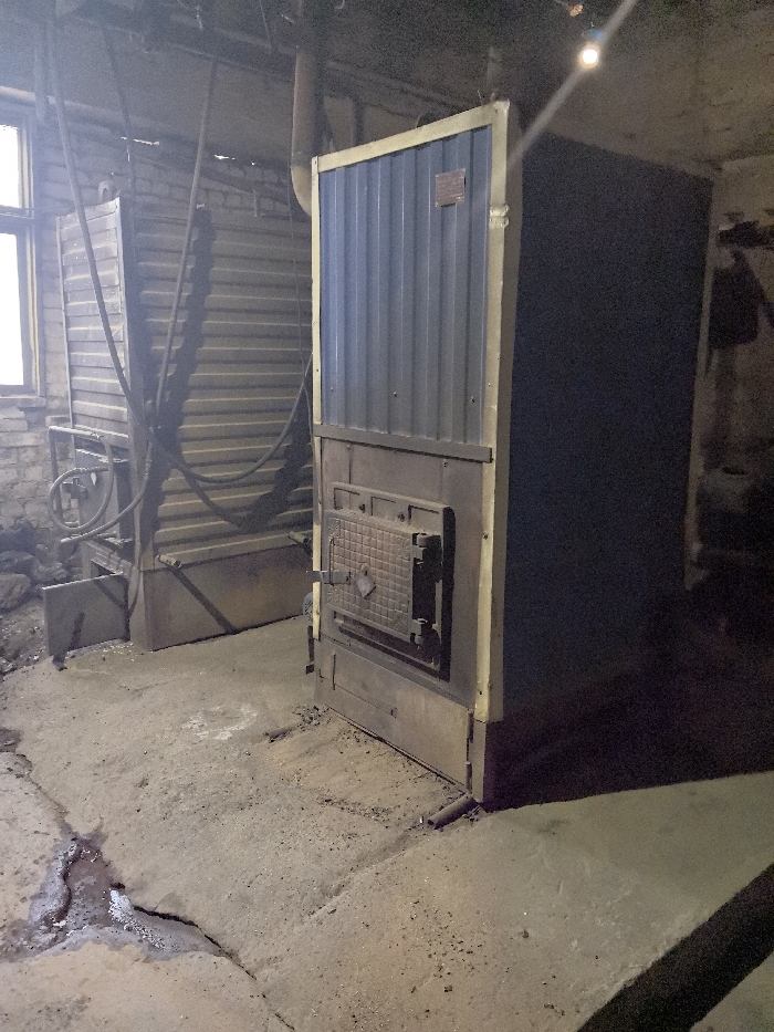The image depicts an interior view of what appears to be a utility or boiler room in Ablatuysky Bor, Russia. In the foreground and midground, a tall, rectangular industrial heating unit or furnace stands upright. This unit features a ribbed, dark blue-grey upper panel bordered by a lighter, possibly yellow or beige, frame. Below the ribbed panel, a sturdy, cast-iron door with a textured, grated central section is visible. The door has a small, diamond-shaped handle or latch.
To the left and slightly behind this primary unit, another large, horizontal industrial structure is present, characterized by corrugated metal panels and a network of dark pipes and hoses running across its surface. A small, rectangular window with a white frame is situated on the far left, providing ambient light. The wall adjacent to the window appears to be constructed of brick.
The floor is concrete, cracked and uneven, with a significant dark, damp patch containing liquid in the lower-left quadrant. Dust, debris, and scattered particulate matter are visible across the floor. The overhead area reveals pipes, electrical wiring, and a single exposed light bulb casting a diagonal beam of light towards the right side of the image. The overall environment appears aged, functional, and sparsely lit. There are no individuals present in the scene.