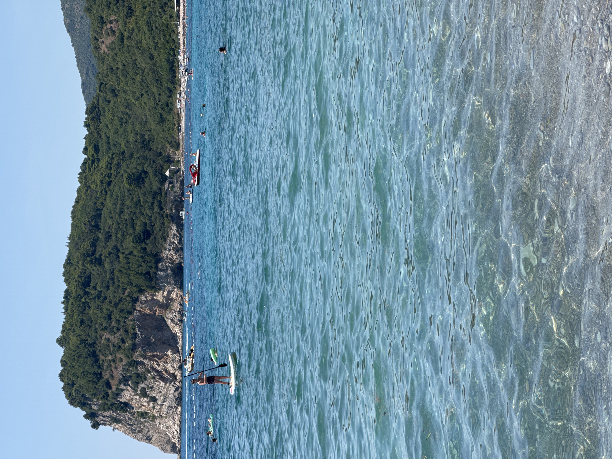 The image depicts a scenic view of the Adriatic Sea near the city of Đurovići, Montenegro. In the foreground, the clear turquoise water reveals the pebble seabed beneath. Several individuals are engaged in water activities. A person stands on a paddleboard, holding a paddle, while another person is seen on a paddleboard further in the distance. Other people are swimming or relaxing in the water. In the mid-ground, a small red pedal boat with occupants navigates the sea. The background features a lush green, heavily wooded cliff face rising from the sea, characteristic of the Montenegrin coast. A sandy beach with umbrellas and more people can be seen along the shoreline at the base of the cliffs, indicating a popular tourist destination. The sky is clear and blue.