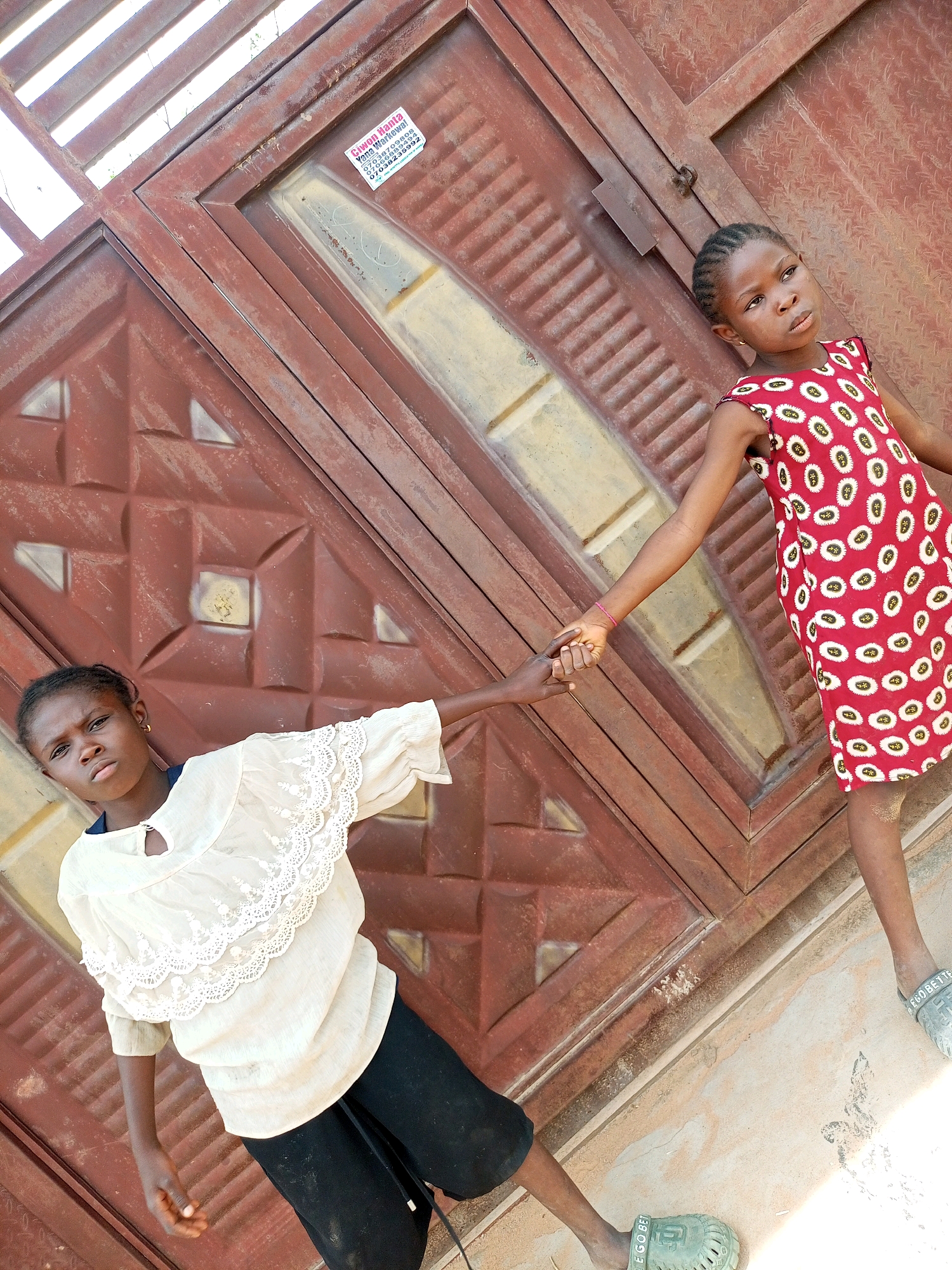 Two young girls stand in front of a brown metal gate. The girl on the right wears a red dress with white and yellow circular patterns. The girl on the left wears a white lace-trimmed blouse and black shorts. They are holding hands. The gate has a diamond pattern and some vertical slats at the top. A sticker with writing in what appears to be a foreign language is affixed to the gate. The ground in front of the gate is dusty and sandy. The scene appears to be outdoors, likely in a residential area in Tempure, Nigeria.