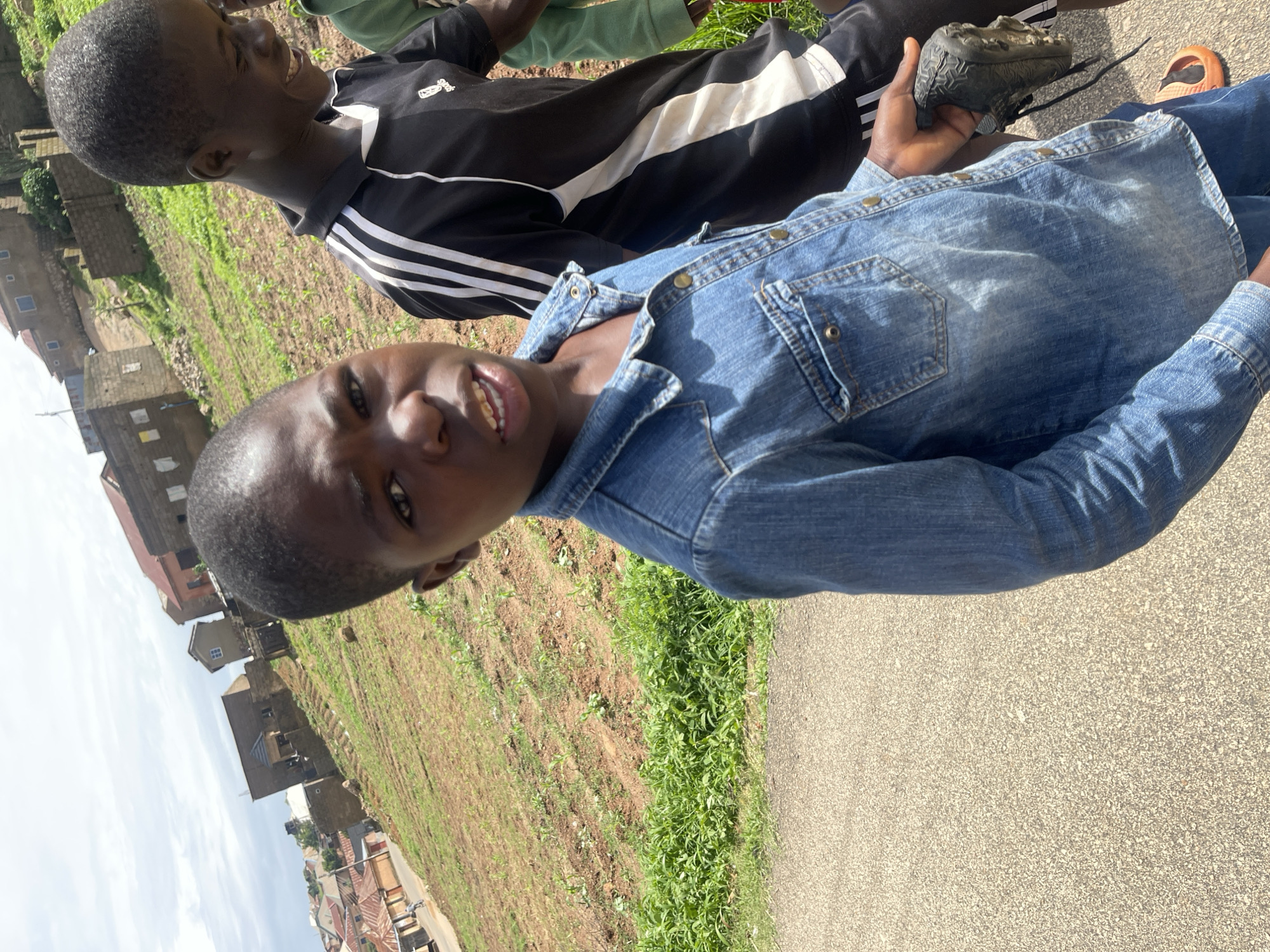 In an outdoor scene in Jos, Nigeria, two young boys are visible, with the foreground dominated by one boy in a denim shirt. The boy in the foreground, facing the camera, is looking directly at it with a slight grimace, his mouth open and teeth visible. He is wearing a blue denim shirt with brass buttons and chest pockets, and appears to be holding a black shoe in his right hand. Behind him, another boy is partially visible, smiling and looking to his right. He wears a black and white striped athletic shirt. The environment suggests a hillside setting with scattered residential buildings and patches of tilled earth and green vegetation. The sky is overcast. The scene appears to be on a paved path.