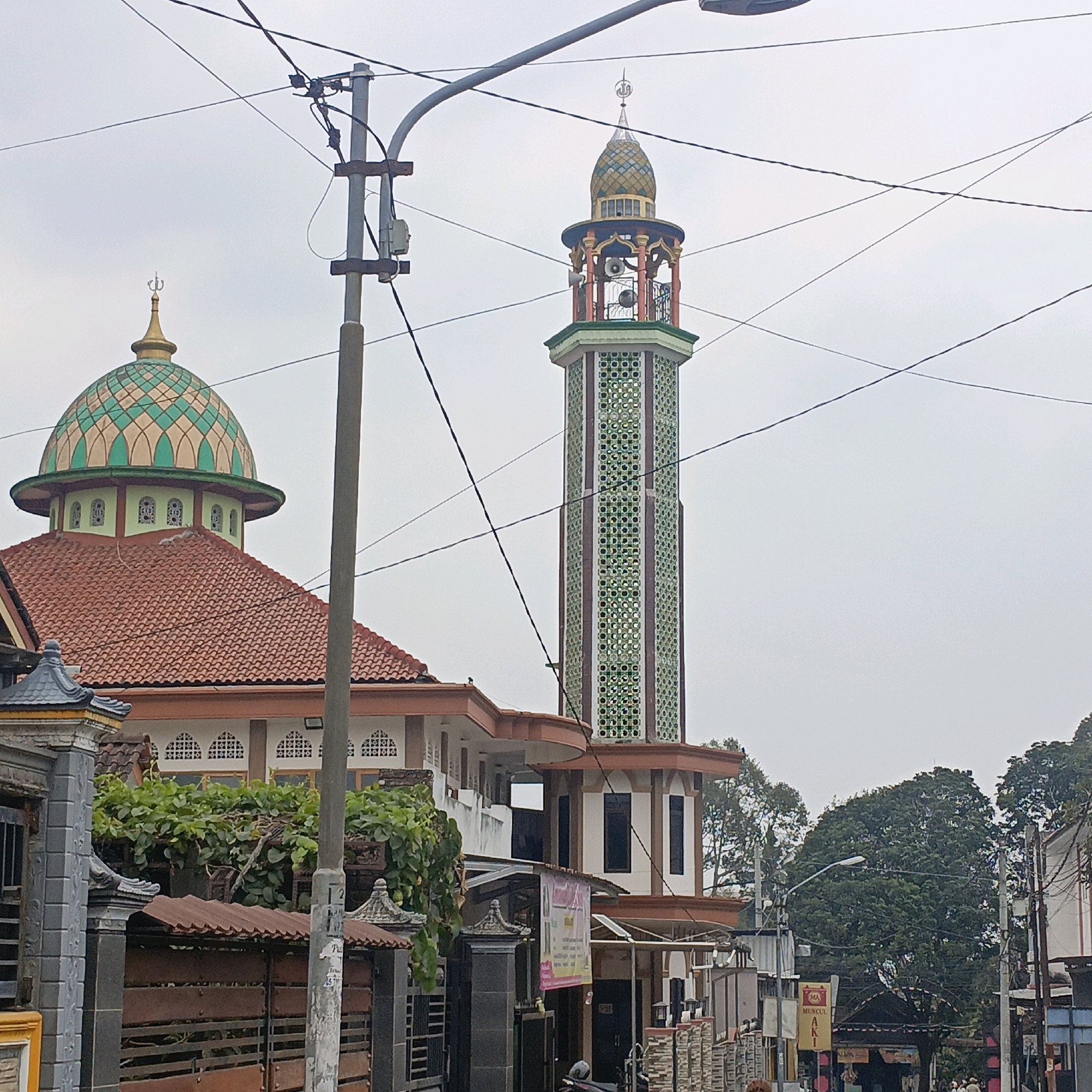 The image shows a mosque in Salatiga, Indonesia, with a tall, octagonal minaret decorated with green geometric patterns and a tiered, colorful dome. The main building of the mosque has a red tiled roof and white walls with arched windows. In the foreground, a utility pole with numerous wires is prominently placed, partially obscuring the mosque. Behind the mosque, lush green trees are visible, and the sky is overcast. The scene depicts a typical street view in a Southeast Asian city, with the mosque serving as a focal point.