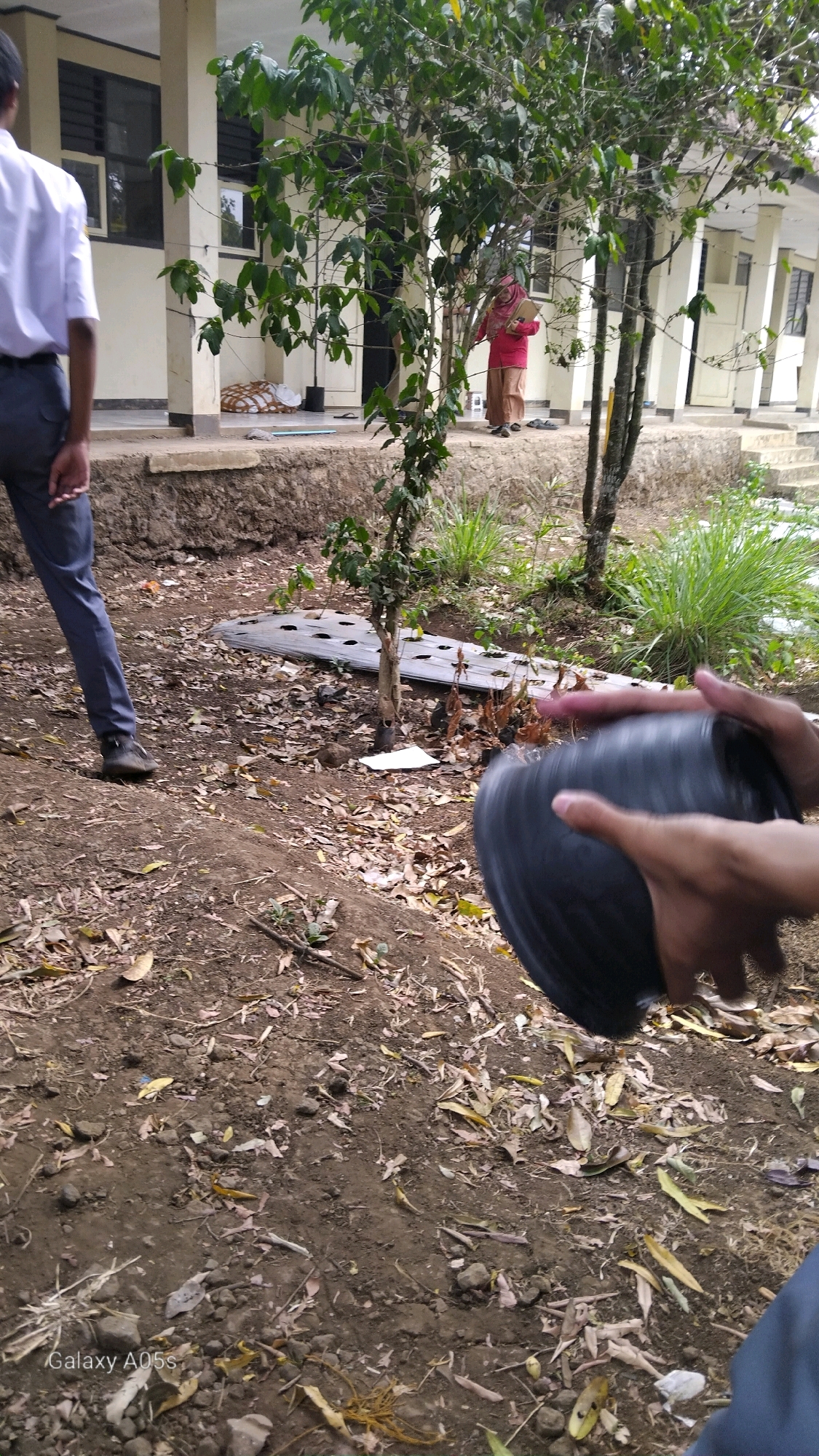 The image shows an outdoor scene, likely on the grounds of a school or institution, with a building in the background. In the foreground, a person's hands are holding a black, ribbed cylindrical object, possibly a pot or a basket. Another person, wearing a white shirt and blue pants, is partially visible on the left side of the frame, facing away from the camera. A woman in a pink shirt and headscarf stands near the building in the background. The ground is covered with soil, fallen leaves, and some scattered debris, including a piece of black plastic sheeting with holes. There are trees and green foliage present in the environment. The scene appears to be in Sumedang, Indonesia, as indicated by the "Galaxy A05s" watermark, a common feature on photos taken with Samsung mobile devices.