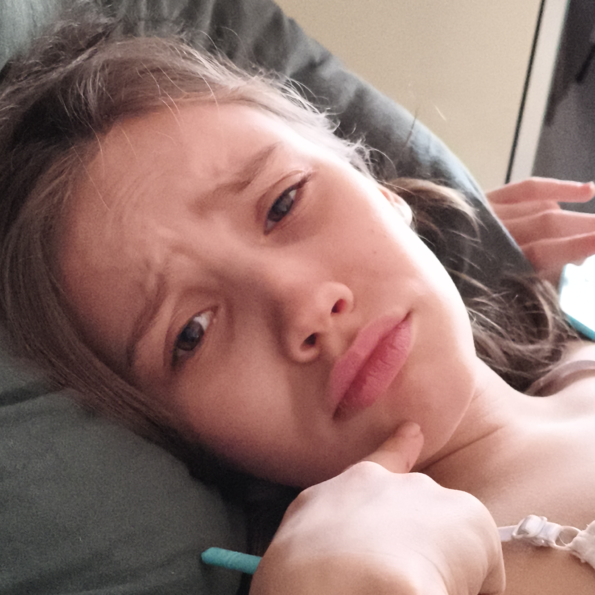A young person with light brown hair is lying on a green couch. The person has a hand raised to their chin and is looking at the camera with a questioning expression. They are wearing a white undergarment. A teal object is visible in the lower left corner. The location is unspecified.