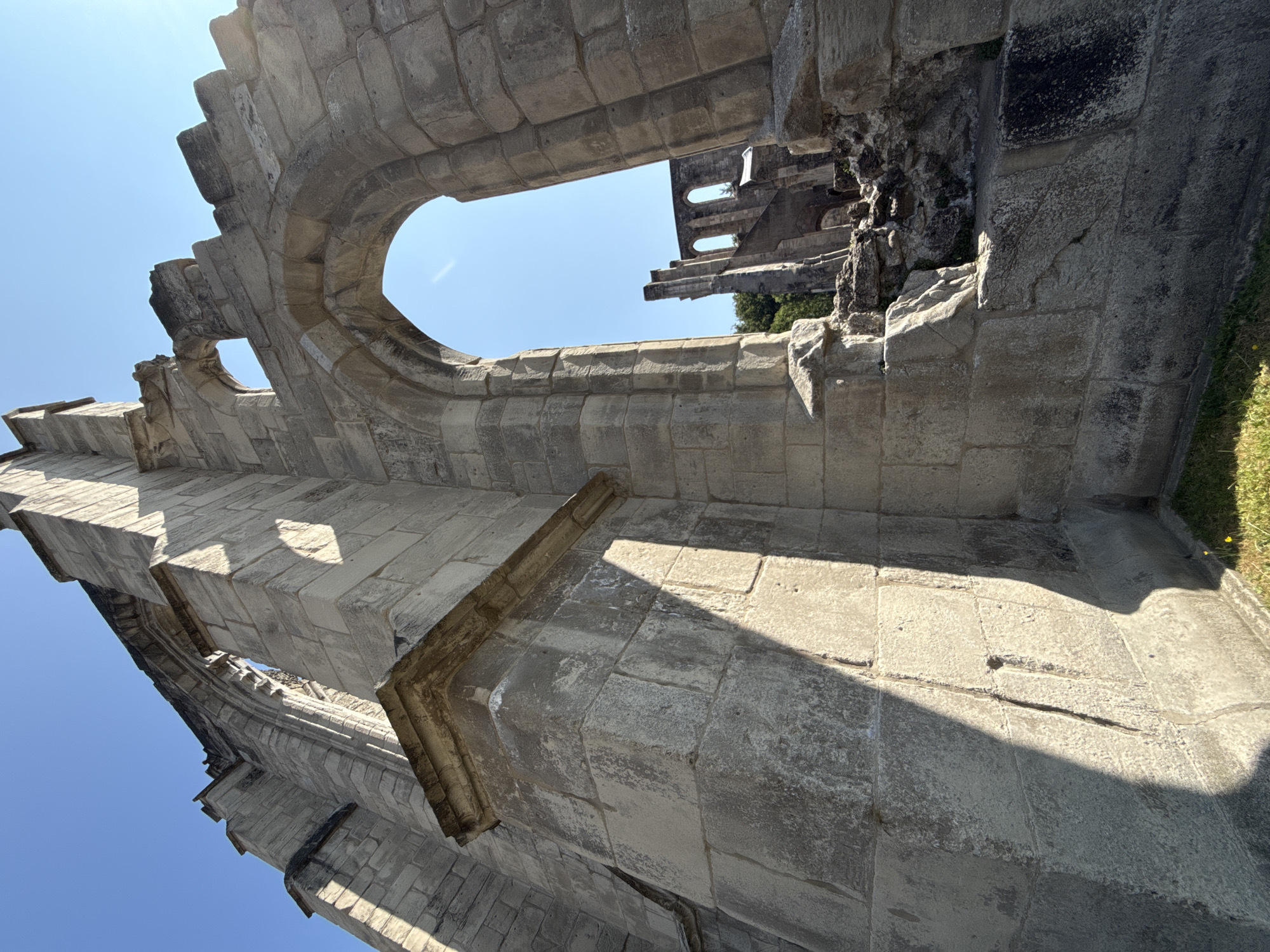 The image depicts the exterior of the Walkenried Abbey ruins in Walkenried, Germany, under a clear blue sky. The perspective is low and looking upwards at the weathered stone architecture. Large, rough-hewn stone blocks form the walls, with evidence of decay and missing sections. A prominent, arched opening is visible in the foreground, framing a view of more ruined structures and trees in the distance. Sunlight casts strong shadows across the stone surfaces, highlighting the textures and age of the building. The ground is covered with grass, with a few small yellow flowers visible. There are no people or animals present in the image.