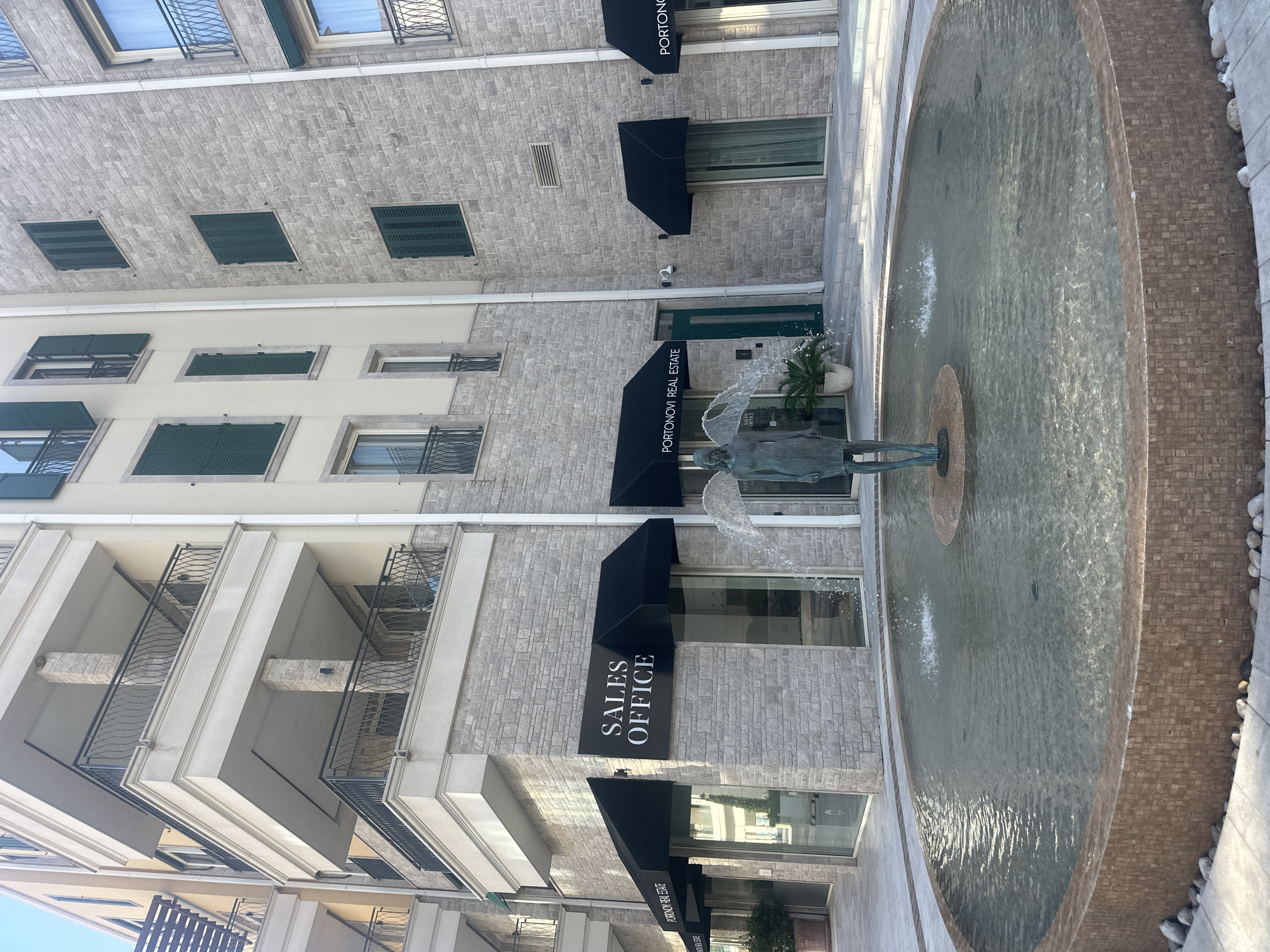 The image shows a fountain in the foreground with a statue of a winged figure in its center. The fountain is circular and tiled with small, mosaic-like pieces in shades of brown. Water is spraying from the statue, creating a splashing effect. In the background, there is a modern building with stone and cream-colored facades. The building has multiple balconies with black railings and green shutters on the windows. There are awnings over storefronts with signs that read "SALES OFFICE" and "PORTONOVI REAL ESTATE." The location is identified as Kumbor, Montenegro. The scene appears to be a public space, possibly a plaza or courtyard, adjacent to commercial or residential properties. There are no people visible in the image.