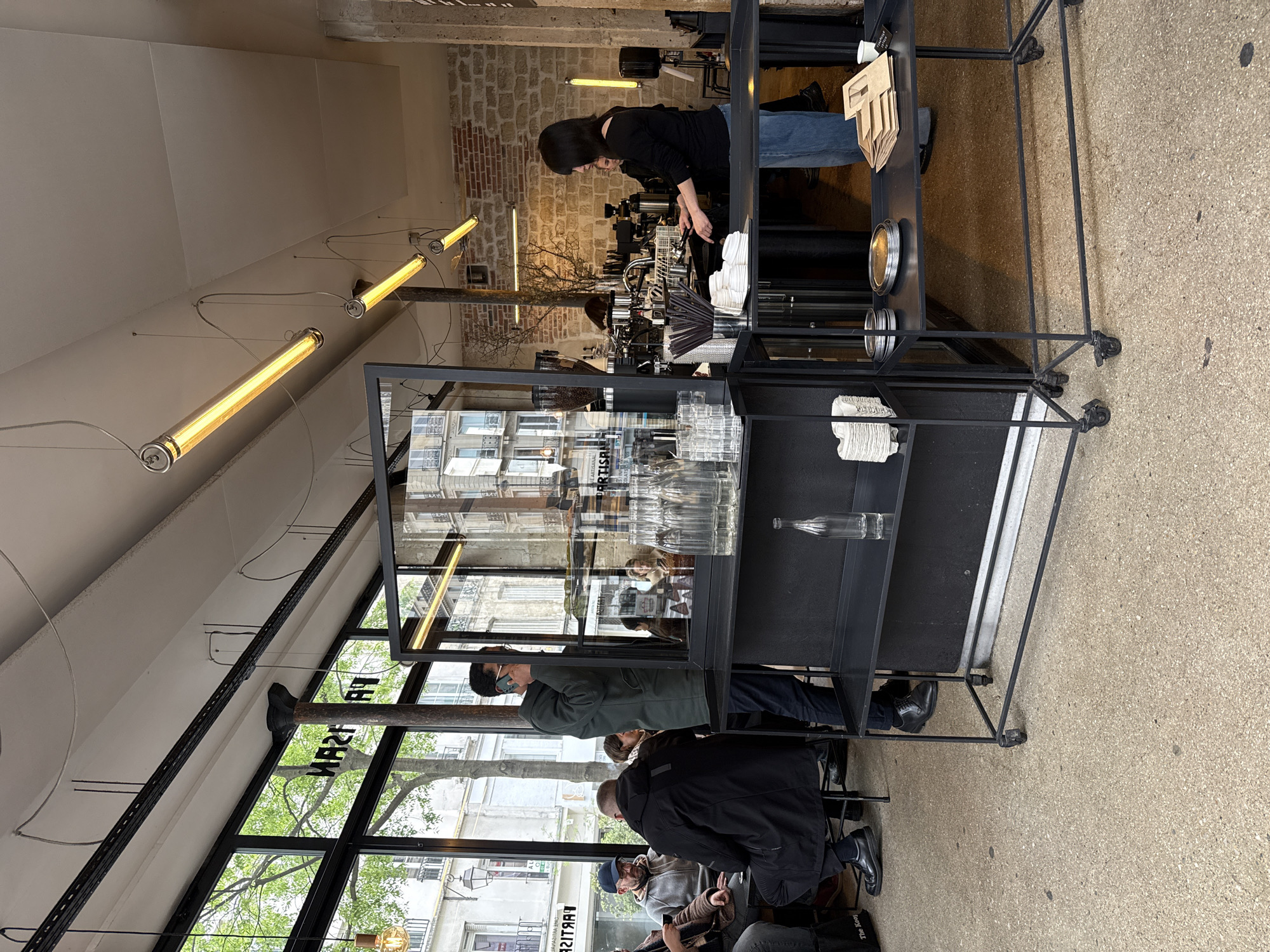 The image depicts the interior of a cafe in Paris, France. A barista is preparing drinks behind a counter, facing away from the camera. The cafe has large windows overlooking a street lined with buildings and trees. Customers are seated at tables near the windows. A black metal shelving unit stands between the customers and the barista, containing glassware, napkins, and other cafe supplies. Linear tube lights hang from the ceiling.