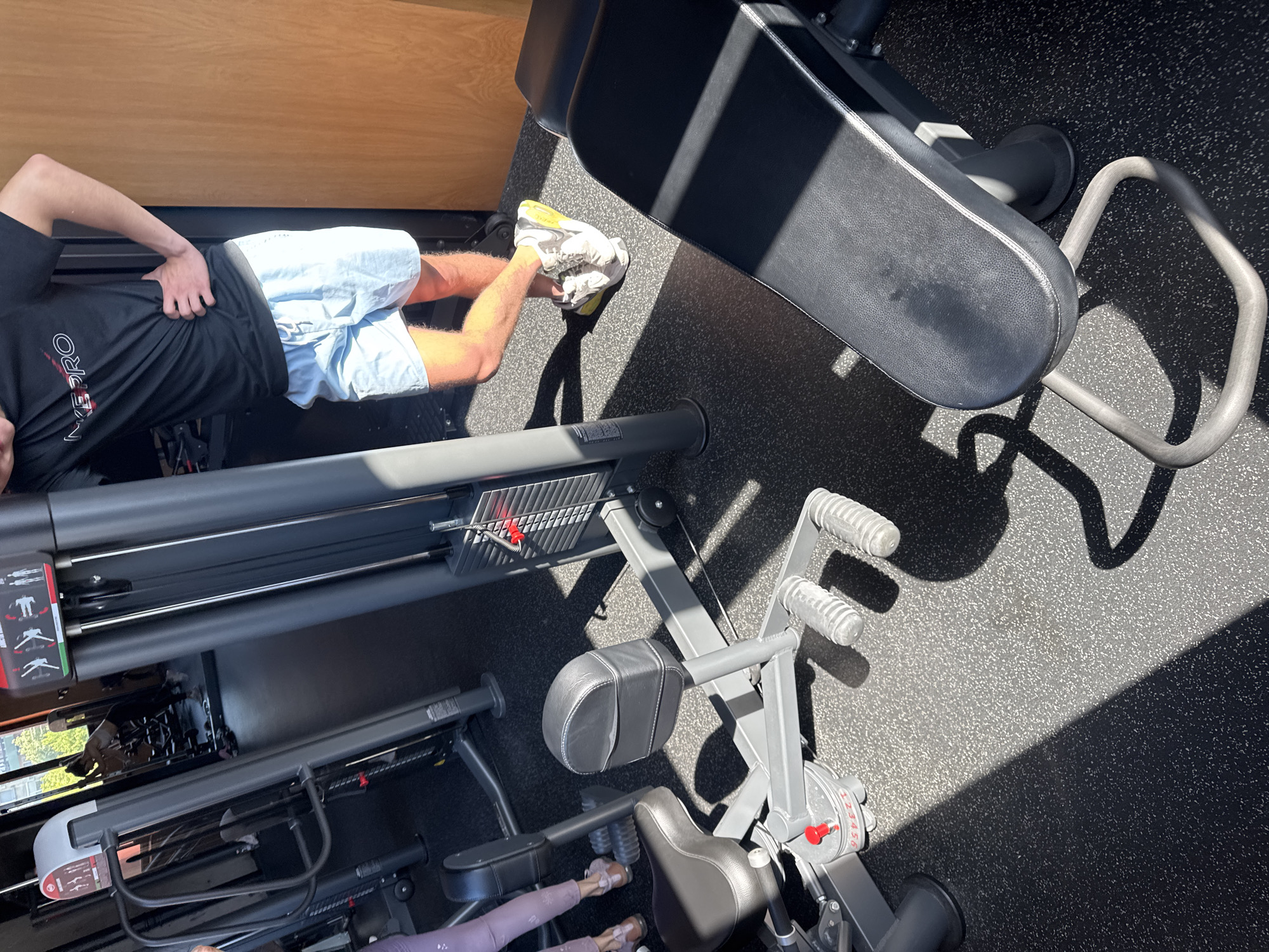A high-angle shot depicts a person lying on a leg curl machine in what appears to be a gym. The person is wearing a black t-shirt with red lettering, light blue shorts, and white sneakers with yellow accents. The leg curl machine has a black padded seat and multiple adjustable components in gray metal. The surrounding floor is black with a textured surface. Light streams in, creating strong shadows across the gym equipment and floor. The location is not identifiable as Plovdiv, Bulgaria.
