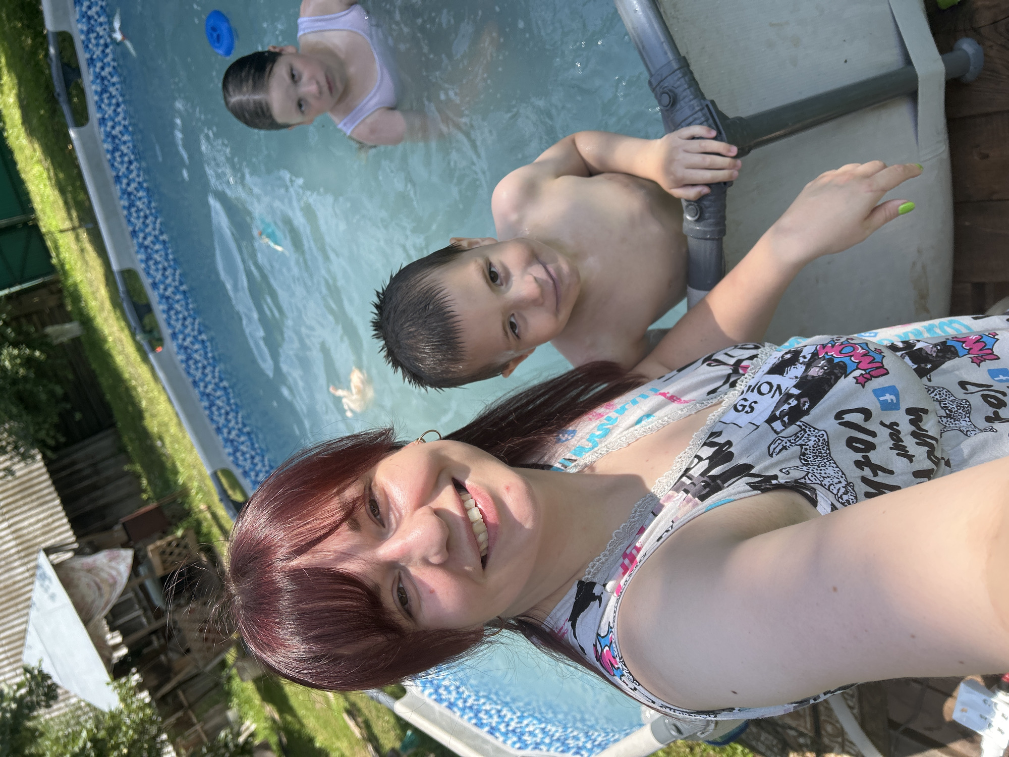 A medium shot captures a woman with reddish-brown hair and bangs, smiling at the camera. She is wearing a black and white patterned dress with pink and blue accents and has a gold hoop earring. She is in a swimming pool with clear blue water. To her right and slightly behind her is a young boy with dark, wet hair. He is shirtless and looking at the camera with a slight smile. Further back in the pool, a young girl with dark, wet hair is partially visible. She is wearing a white tank top and looking at the camera with a stern expression. A blue ring float is visible in the water behind the girl. The background shows a wooden deck with a grey border around the pool, and beyond that, a grassy area with trees and a rustic wooden structure, possibly a shed or gazebo. The overall scene depicts a family enjoying a summer day in a backyard swimming pool.
