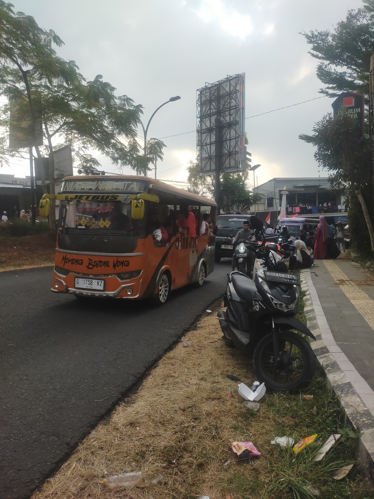 The image depicts a street scene in Tuwel, Indonesia, with a prominent orange tourist bus on the left, its side open with passengers visible. The bus has text in Indonesian on its front, including "MOMONG BOJONE WONG" and a license plate "G 1758 KZ". To the right of the bus, parked along a sidewalk, are several motorcycles, one of which is a black Honda scooter with a license plate "D 4458 ZFE". People, some wearing hijabs, are gathered on the sidewalk, and there is a large billboard in the background. The environment appears to be a busy street with trees, overhead power lines, and buildings lining the road. Litter is visible on the dry grass along the roadside.