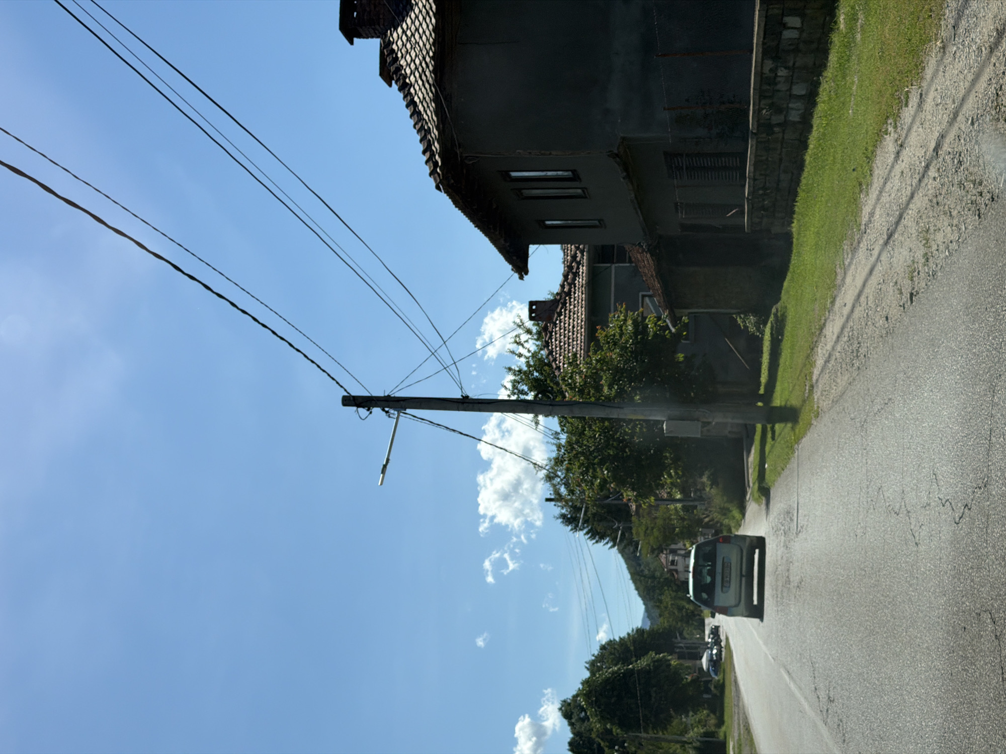 In Beli Osam, Bulgaria, a light blue car travels down a paved road on a sunny day. The car is in the right lane, moving away from the viewer, with trees lining the left side of the road and houses and more trees on the right. The sky is a clear blue with a few white clouds. Power lines are visible overhead, supported by a utility pole on the right side of the road. The road has some cracks and patches. A low stone wall runs along the side of the road in front of a house.