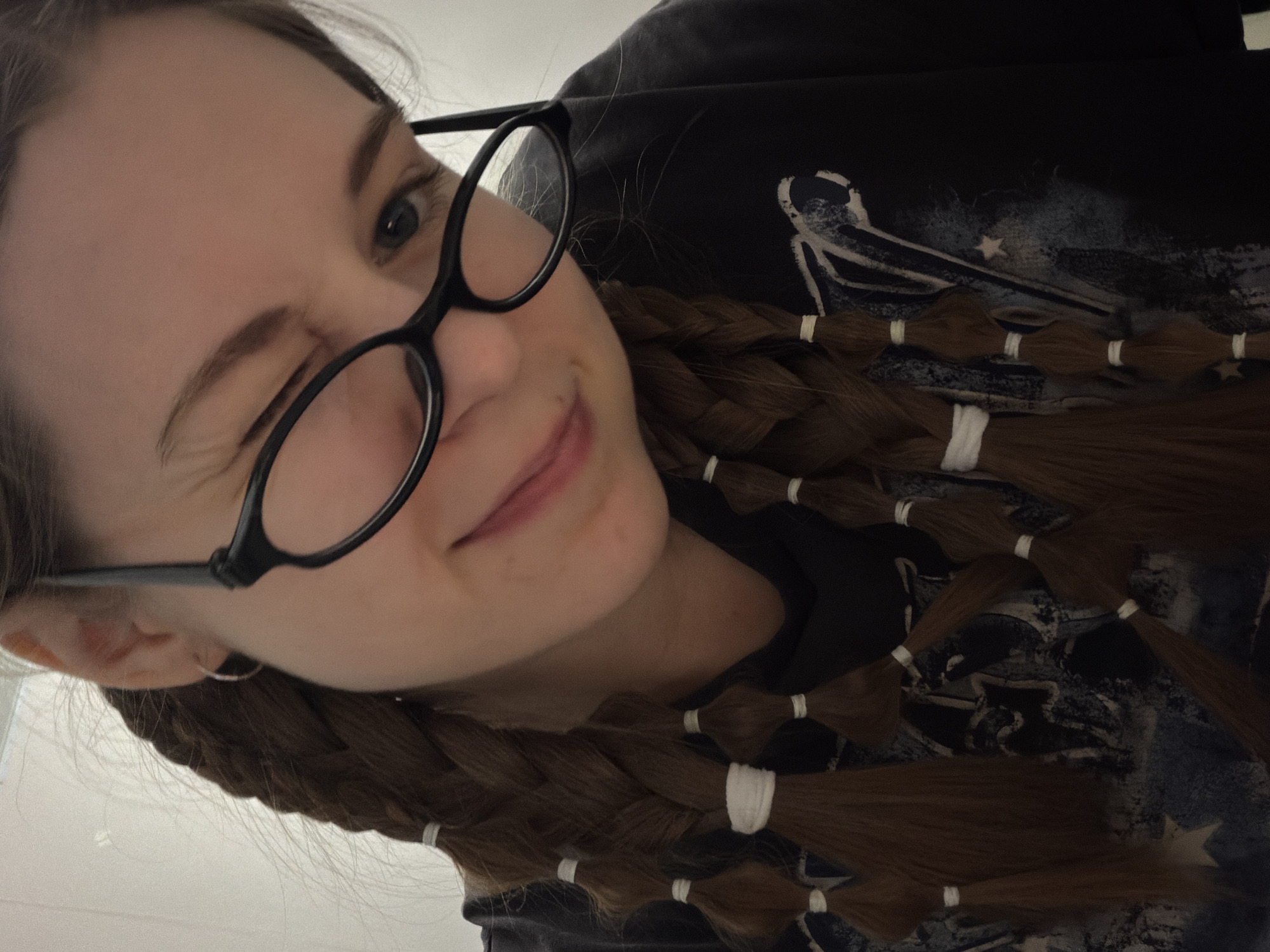 A close-up selfie captures a young woman from the chest up, winking playfully with her left eye and displaying a slight smile. She wears black-framed glasses, which are prominently featured on her face. Her left eye is closed in a wink, causing a visible crinkle at the corner, while her right eye is open and looks towards the camera. A small silver hoop earring is visible on her left earlobe.
Her long, brown hair is styled in multiple segmented braids, resembling bubble braids, secured at regular intervals with numerous small white elastic hair ties. These braids are draped over her shoulders and chest, partially covering a dark-colored garment, likely a hoodie or t-shirt. The garment features an abstract graphic design with white and grey elements that could depict a landscape or celestial scene, including what appear to be faint star-like shapes. The background is a plain, light, indeterminate indoor surface. The image is taken from a slightly low angle, giving the perspective of looking up at the subject, and is oriented diagonally. No text is visible in the image. The scene is set in Perm, Russia.