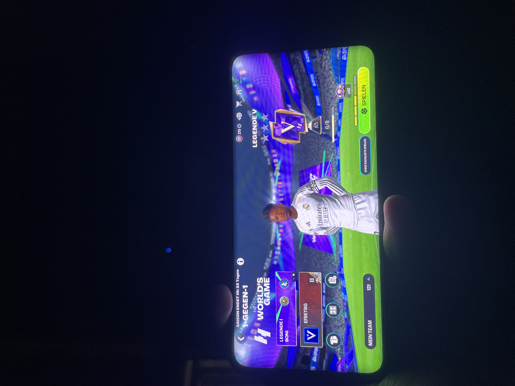 The image shows a smartphone screen displaying the video game FIFA Mobile. The game is in German, with the title "1-GEGEN-1" and "THE WORLD'S GAME". The player's rank is "LEGENDE V" with three stars, and they have completed 6 out of 8 stages. A player character, wearing a Real Madrid jersey with "Emirates FLY BETTER" printed on it, is visible on the right side of the screen. The bottom of the screen shows options like "MEIN TEAM" (My Team) and a button to start a game, "SPIELEN" (Play). The phone is held in a hand, obscuring part of the bottom left corner of the screen. The background is dark, suggesting the image was taken in low light conditions.