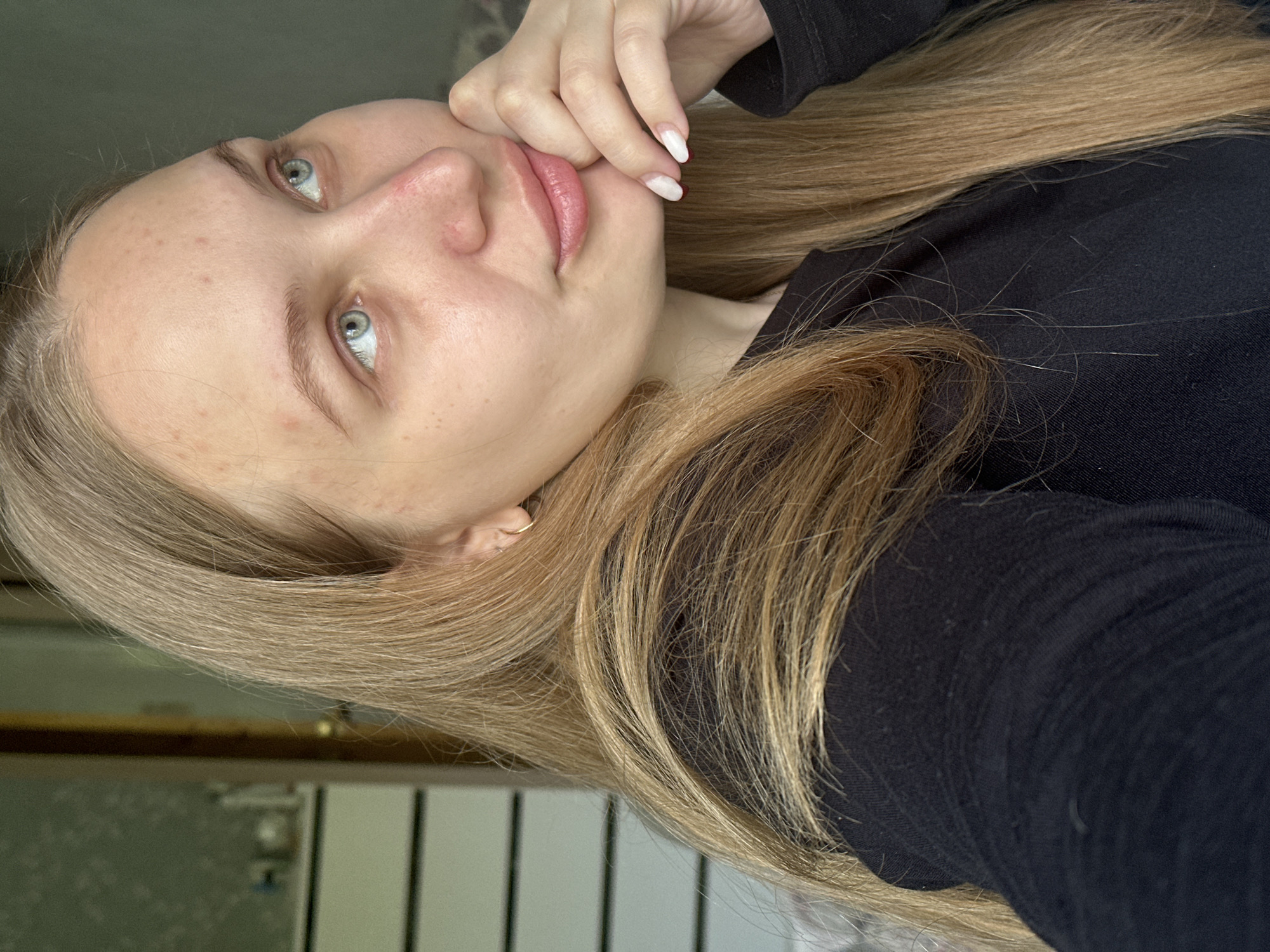A young woman with light blonde hair and blue eyes is posing for a selfie indoors. She is wearing a black top and has a gold hoop earring in her left ear. Her left hand is resting near her lips. The background is partially obscured, but a white and brown door is visible. The location is Chaikovsky, Russia.