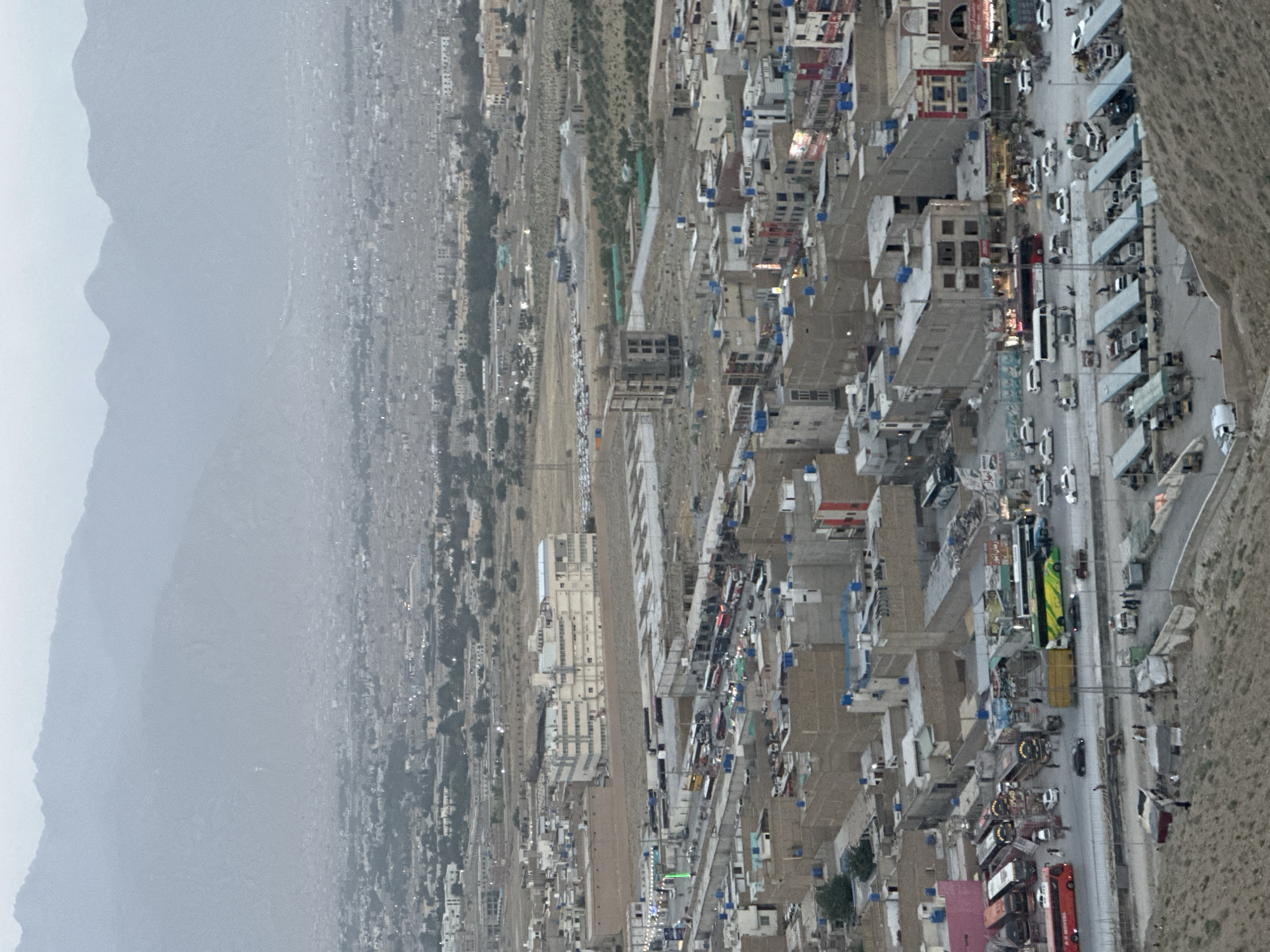 The image shows an aerial view of a densely populated area in Quetta City Tehsil, Pakistan. A mountain range is visible on the left side of the frame, obscured by haze. The city sprawl extends from the mountains towards the right, with buildings of various sizes and designs clustered together. A primary road runs through the center of the image, with a high volume of traffic including cars, buses, and other vehicles. Adjacent to the road are rows of buildings, some appearing to be residential while others are commercial establishments. The scene seems to capture a typical urban landscape with a mix of residential and commercial activities. The overall environment has a somewhat dry and dusty appearance, indicative of the region's climate.