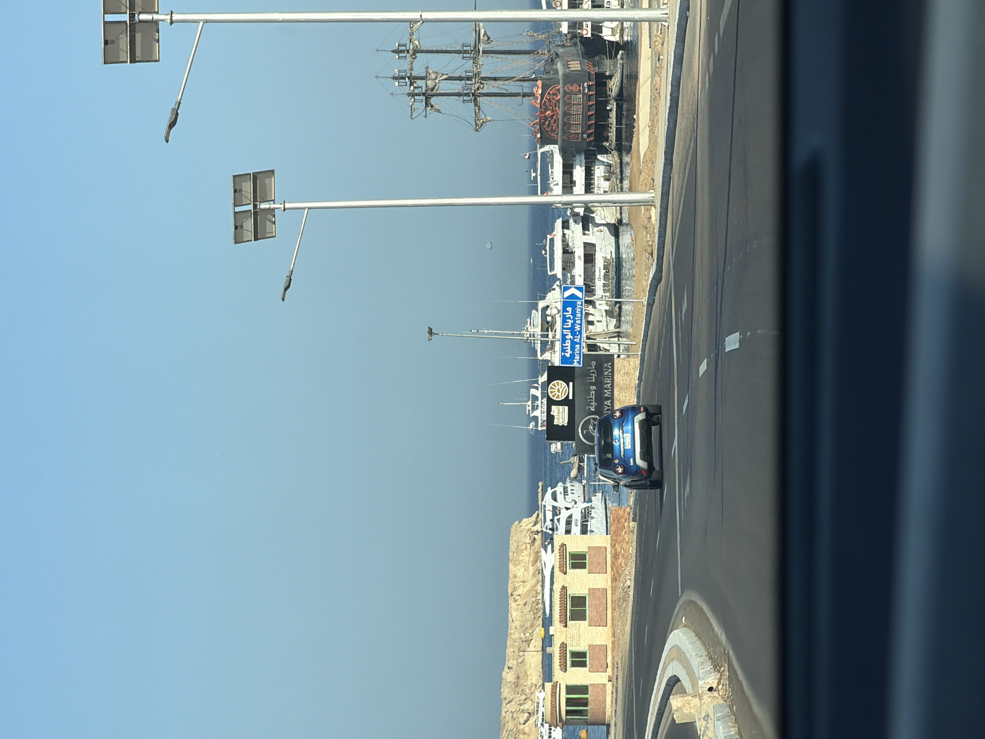 The image depicts a view from a vehicle traveling on a paved road in Sharm El Sheikh, Egypt. To the left of the road, there is a tan-colored building with red brick accents and multiple windows, situated near the water. Beyond the building and extending into the blue sea, numerous boats and yachts are docked, including a large, dark-colored pirate ship with tall masts on the right side of the frame. A blue road sign with white Arabic and English text, indicating "Marina Al-Wataniya," is visible on the right, pointing towards the marina. Two streetlights with solar panels are positioned along the road. The sky is clear and blue, and the overall environment suggests a sunny day at a coastal location. A blue car is driving ahead on the road. The foreground shows the interior of a car's dashboard and windshield.