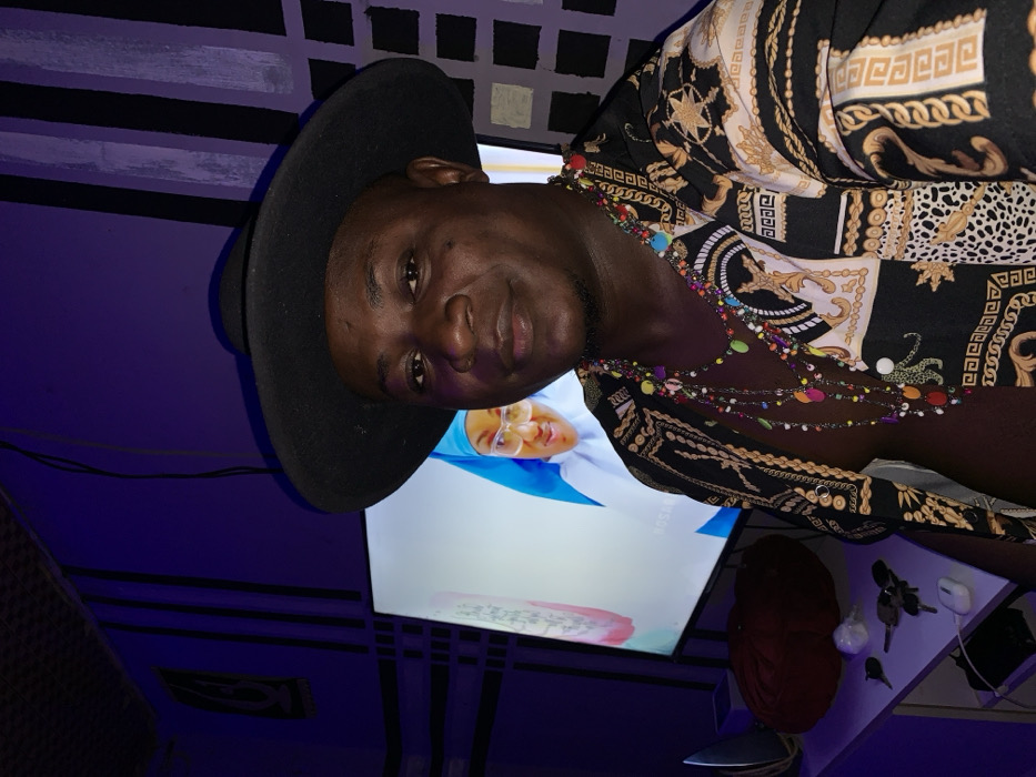 A man wearing a black cowboy hat and a patterned shirt stands indoors, possibly in a nightclub setting with blue lighting. He sports colorful beaded necklaces. Behind him, a television screen displays a woman wearing a hijab. A table cluttered with keys and other objects is visible to the side. The scene has a casual, relaxed vibe.