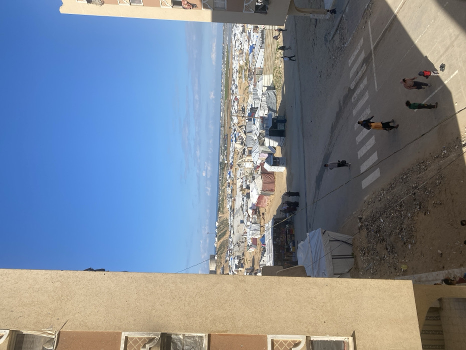 The image presents an elevated, wide-angle view of a densely populated area in Khan Yunis, Palestinian Territory, under a clear blue sky with scattered white clouds. Two multi-story, light-colored buildings frame the scene; the left structure shows signs of damage or makeshift repairs around its windows and a dark opening near the top, while the right building has fabric draped from an upper window.
In the midground, a paved or unpaved road with a white-striped pedestrian crossing extends horizontally. On this road, several individuals are visible: one person carries another on their shoulders near the crossing, while a small group including a child in red stands nearby. Further left, a single individual in a white shirt carries a dark bag while walking. To the left of the road, a mound of dirt and debris is adjacent to a white tent structure. Across the road, several makeshift market stalls, displaying various goods, are set up, surrounded by groups of people.
The background is dominated by a vast encampment comprising hundreds of white and brown tents and various temporary shelters, sprawling across a dusty or sandy landscape. Elevated terrain, possibly hills, is visible beyond the encampment, topped by a large, cylindrical water tank. The Mediterranean Sea is discernible on the distant horizon. The scene is brightly lit by sunlight, casting distinct shadows.