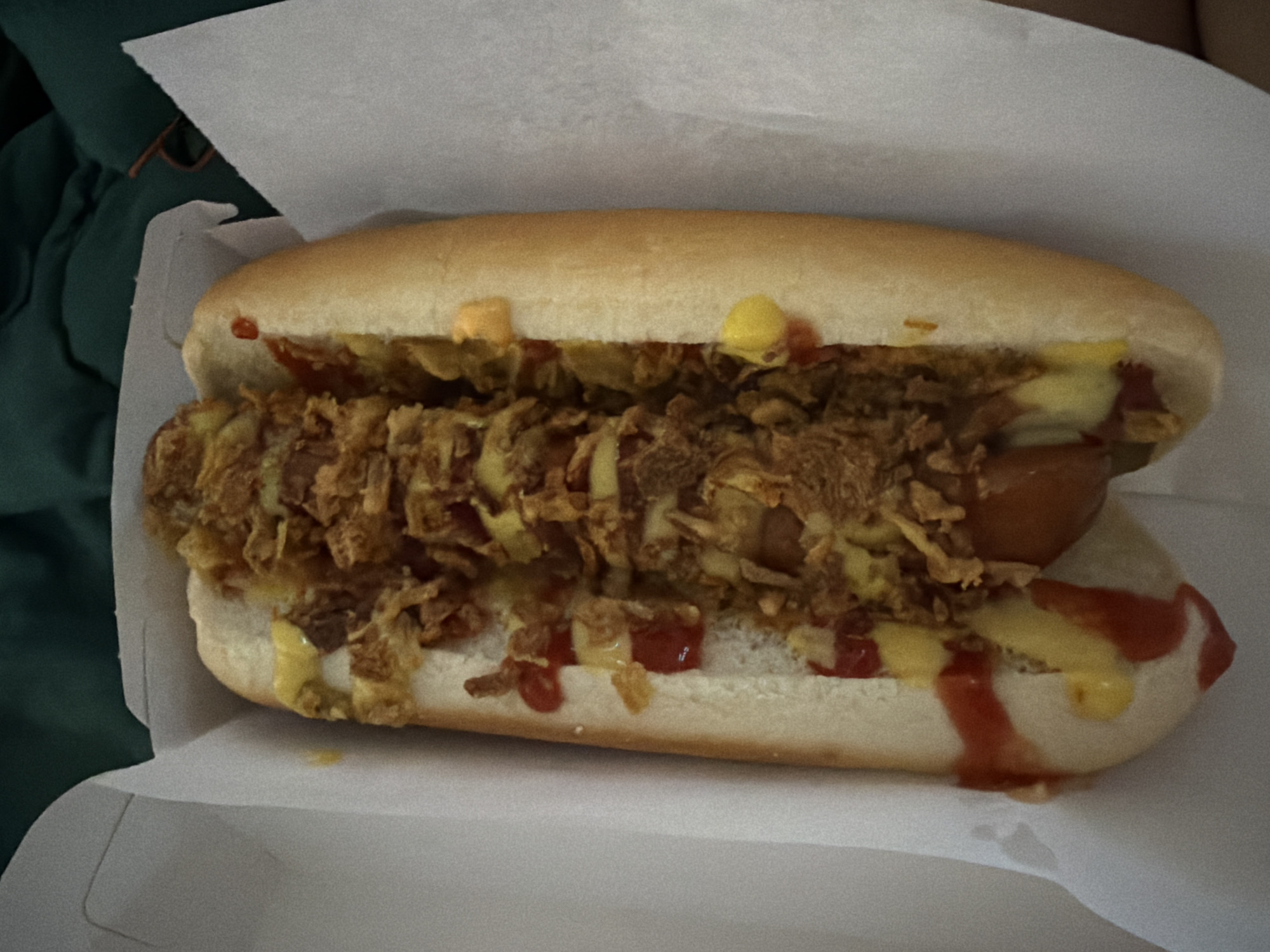 A close-up, top-down shot displays a hot dog in a white paper tray. The hot dog is nestled in a bun and topped with a generous amount of chili, fried onions, mustard, and ketchup. The tray is partially lined with white paper. The background is dark and out of focus, showing hints of dark green fabric, possibly a blanket or clothing. The lighting is artificial and appears to be coming from above and to the left. The image appears to be taken indoors.