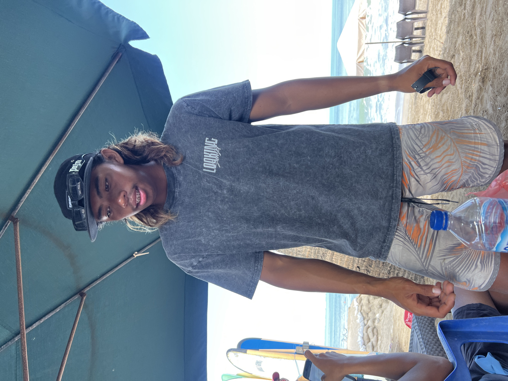 A young male with long, wavy brown hair and a dark baseball cap with sunglasses perched on top stands under a large teal umbrella on a beach. He is wearing a grey, acid-wash t-shirt with the text "LOOKING SUNSET" printed on it in white, and light-colored shorts with a palm leaf pattern in orange and grey. His right hand is extended, holding a black object. In the foreground, a clear plastic water bottle with a blue cap sits on a table to his left, next to a blue plastic chair. Behind him, surfboards are propped up. The beach is sandy, with the ocean visible in the background, and several beach chairs and umbrellas are set up further down the shore. The sky is clear and bright blue. The scene is consistent with a location in Kuta, Indonesia, known for its beaches and surfing culture.
