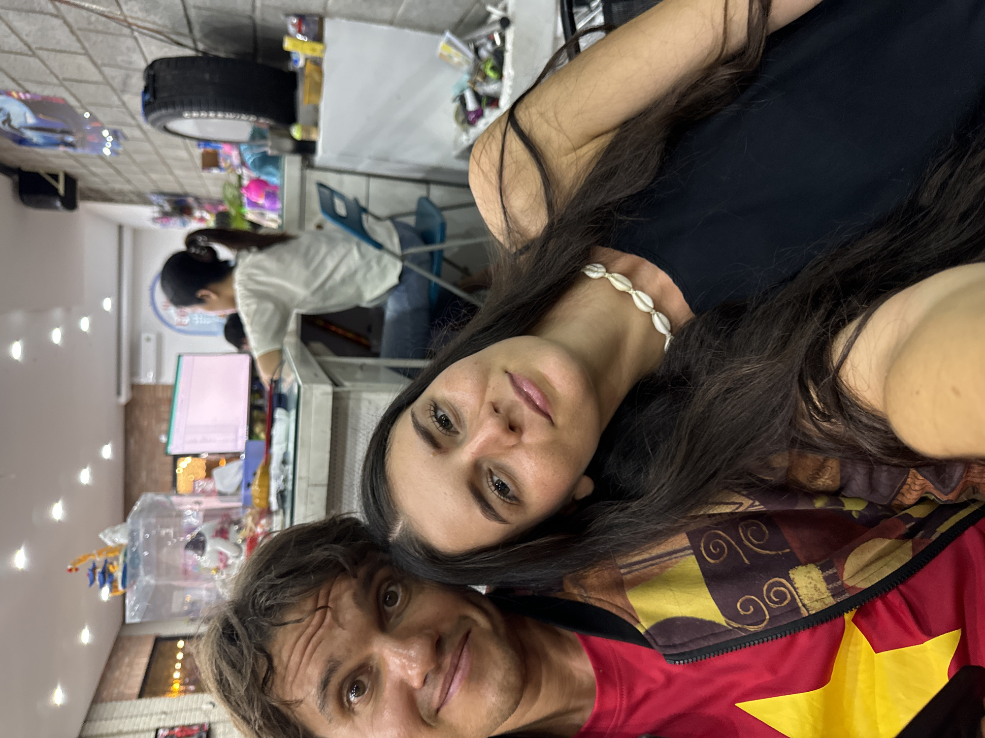 The image, rotated clockwise from its original orientation, captures a selfie of a man and a woman in an indoor retail setting, likely a shop or market in Phu Thuy Ward, Vietnam.
In the foreground, a woman with long, dark hair, wearing a black tank top and a white shell necklace, looks towards the camera with a neutral expression. Beside her, a man with dark, curly hair also looks at the camera with a slight smile. He is dressed in a red jacket prominently featuring a large yellow star on the left side, indicative of the Vietnamese flag, worn over a patterned brown and yellow underlayer.
The background shows a bustling indoor environment, suggesting a shop or convenience store. A third person, a woman with dark hair tied in a ponytail, wearing a white shirt and blue jeans, is visible in the mid-ground, bending over a counter or display area. Shelves laden with various products and merchandise are discernible throughout the background. A car tire is conspicuously mounted on the wall in the upper section of the image. The scene is illuminated by artificial lighting, indicating an indoor setting where the time of day is not apparent. No clearly legible text or signage is visible within the frame. The man's jacket with the Vietnamese flag motif strongly supports the provided location context.