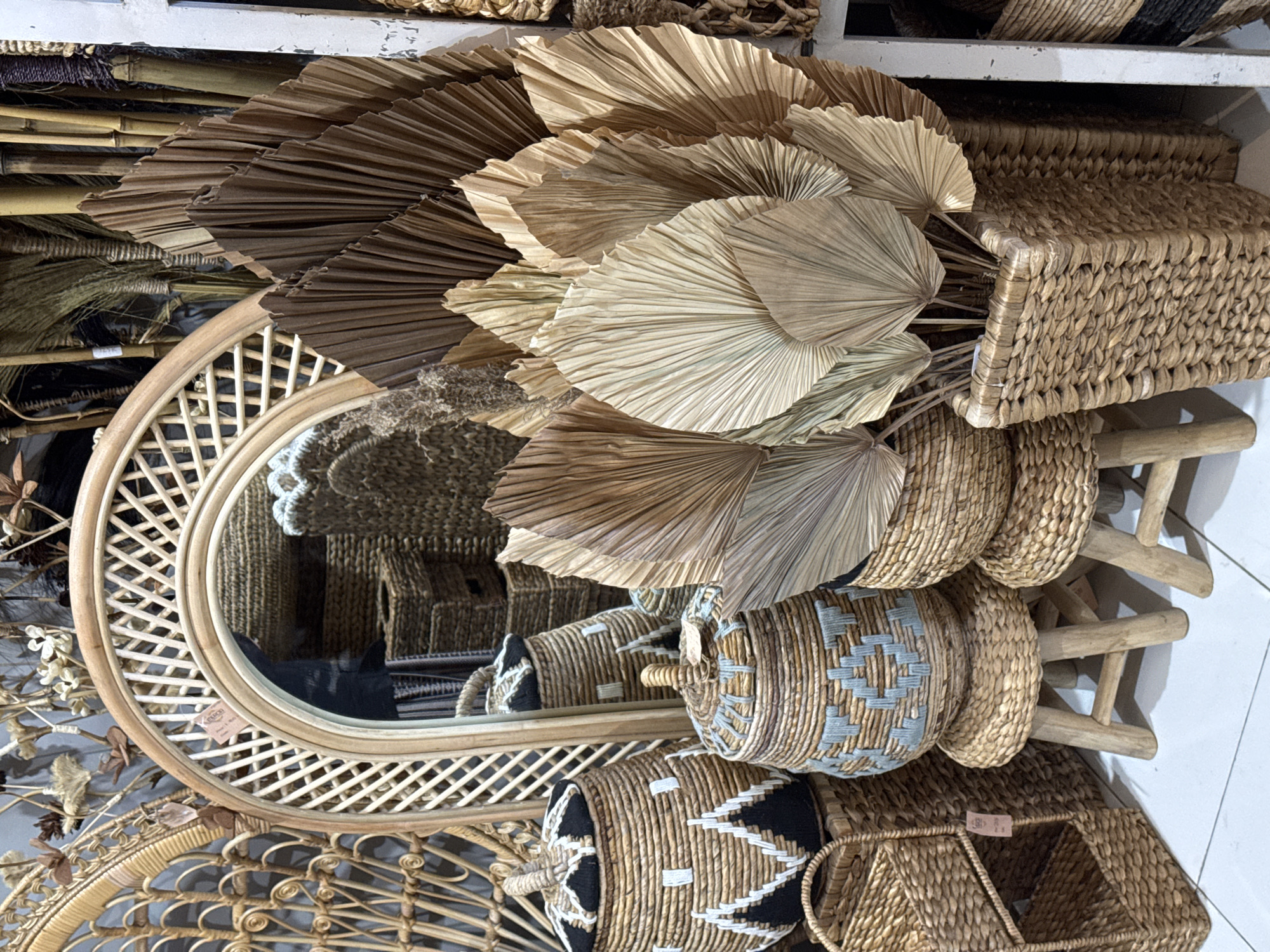 This is a photograph of a retail display of handcrafted woven goods in a store located in Ubud District, Indonesia. The scene is filled with natural materials, predominantly rattan and dried palm leaves. In the foreground, several woven baskets of various sizes and designs are arranged on and around small wooden stools. One large cylindrical basket with a black and white patterned lid sits prominently on the left. To its right, a smaller, similarly shaped basket features a blue and grey woven pattern. In front of these, a two-tiered woven shelf unit holds more baskets.
Dominating the center and right of the image is a large arrangement of dried palm leaves, fanned out in a naturalistic display. These leaves are a range of earthy tones, from light beige to deep brown. They are arranged in a tall, rectangular woven planter. Behind the palm leaves, a tall bamboo structure and various other woven items are visible.
A large, arched mirror with a rattan frame is positioned behind the baskets and to the left of the palm leaf display, reflecting a portion of the store's interior which appears to be stocked with more woven baskets and other decorative items. The overall impression is one of a well-curated display showcasing artisanal craftwork with a natural, bohemian aesthetic. There are no people visible in the image. The floor is tiled in a light color.