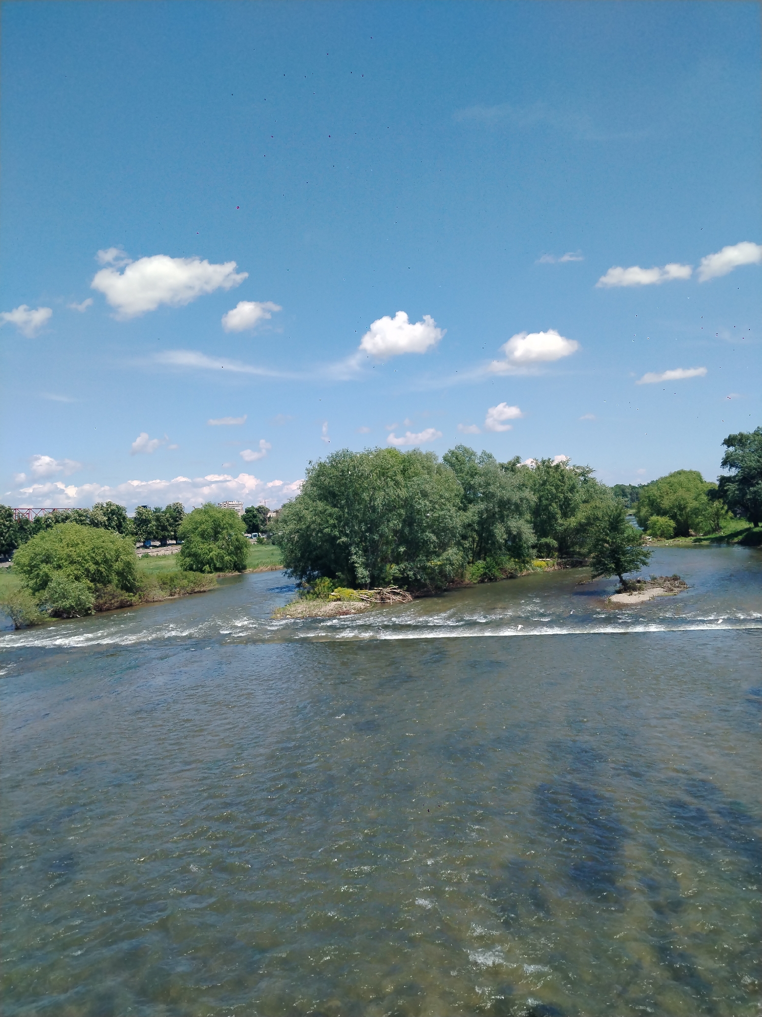 The image depicts a wide river flowing through an urban area in Plovdiv, Bulgaria. The river has a shallow weir or low dam running across it, causing ripples and small rapids. Several small, tree-covered islands are visible in the middle of the river, covered by dense, leafy green trees. In the background, across the riverbank, there are more trees and some buildings indicating a cityscape. The sky is bright blue with scattered white clouds, suggesting a clear, sunny day. The water appears somewhat murky but reflects the light from the sky, giving it a slightly shimmering appearance.