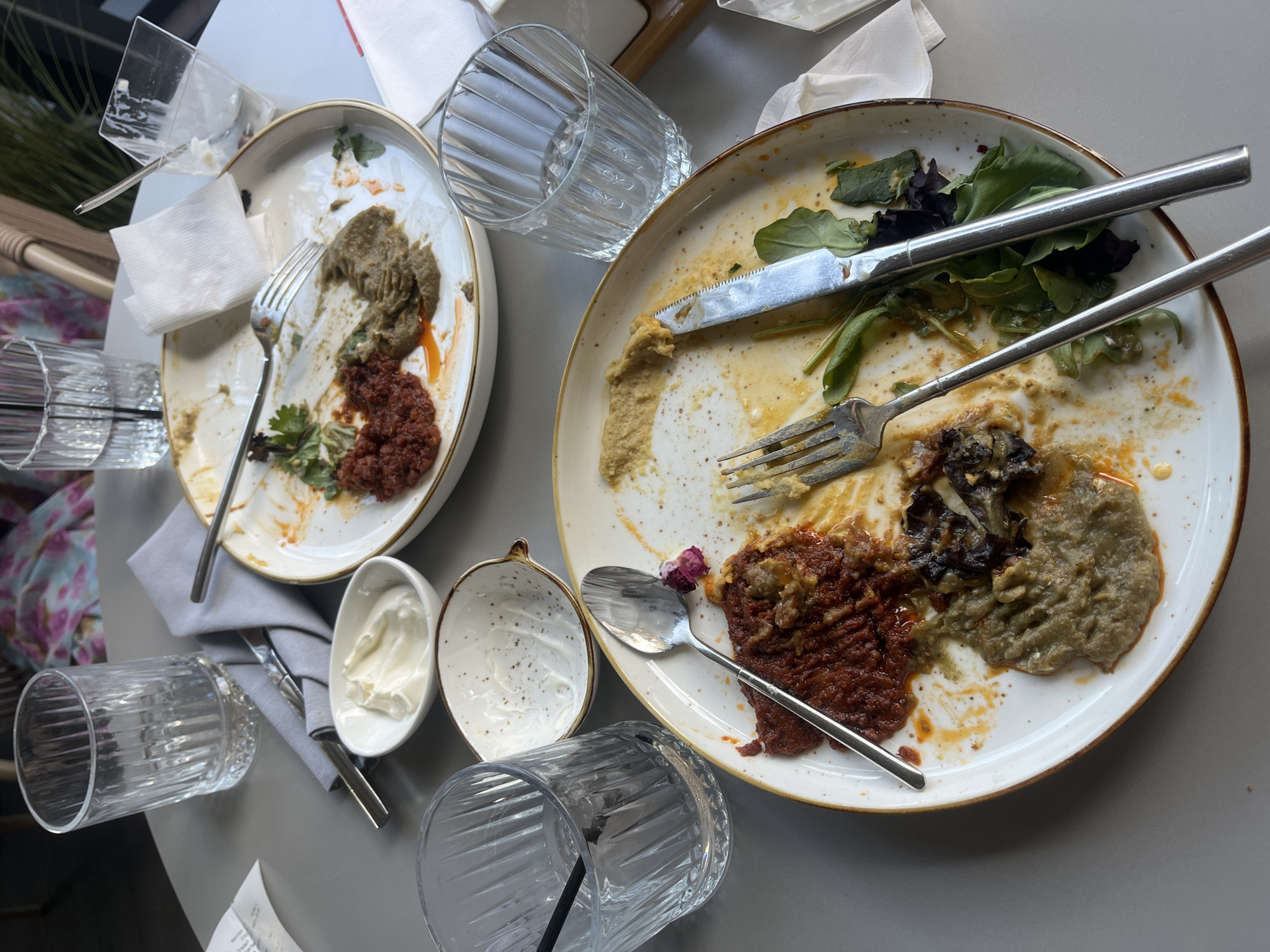 The image depicts a messy table at an outdoor restaurant in Almaty, Kazakhstan, after a meal. Two plates of partially eaten food, likely mezze or appetizers, are visible. One plate contains remnants of a red sauce, a puréed green dish, and some scattered greens. The other plate shows a similar red sauce and green purée, along with some roasted vegetables. Utensils, including forks, a knife, and a spoon, are scattered across the plates and table. Several glasses, some containing water and ice, and a small bowl of what appears to be sour cream or yogurt, are also present. The table is a light gray color, and the background shows blurred elements of outdoor seating and foliage, suggesting a pleasant dining environment. The overall scene conveys the aftermath of a shared meal, with the remaining food and used dishes indicating the meal has concluded.