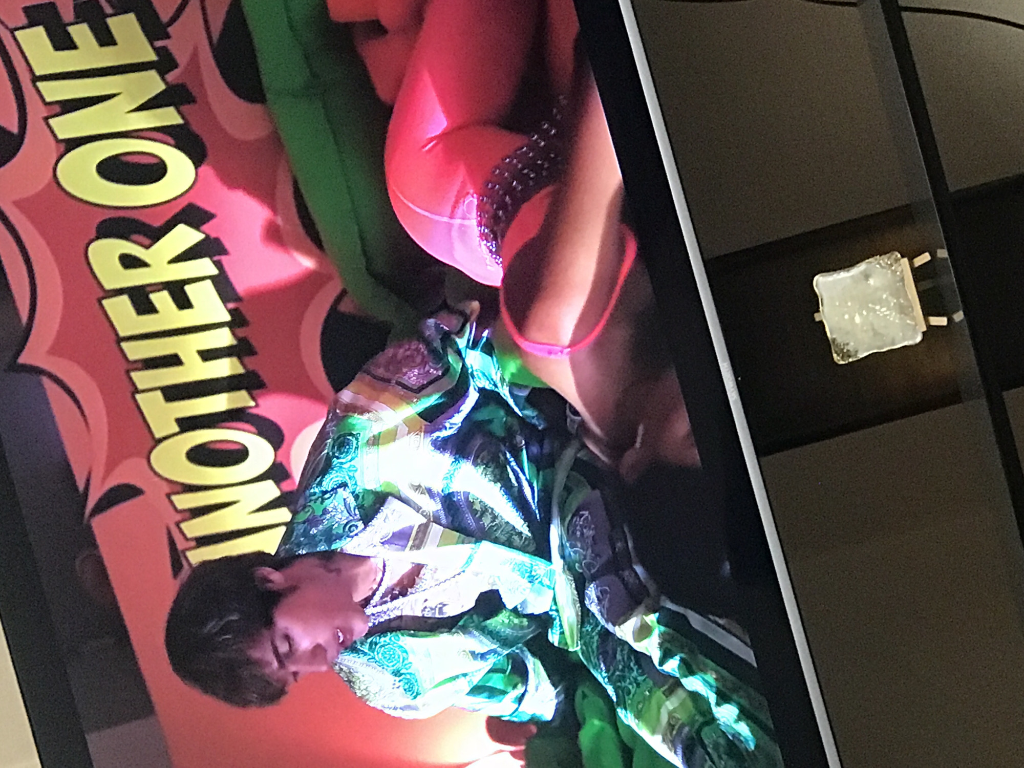 The image shows a television screen displaying a scene from a music video. A performer with dark hair is wearing a brightly colored, patterned robe. Adjacent to this figure is another figure wearing bright pink attire. In the background, a graphic displays the words "ANOTHER ONE" in yellow letters. The image captures the scene as displayed on a modern flat-screen television set within a likely domestic setting. No information is present to suggest a location in Yerevan, Armenia. A small, decorative object is visible on a dark, glossy surface next to the television.