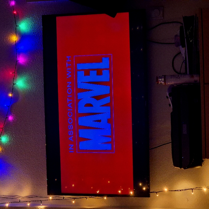 The scene is an interior view within a residential setting in Strasbourg, France, depicting a dimly lit room focused on a television screen. A flat-screen television with a black bezel is mounted on a textured, light-colored wall. The screen displays a vibrant red background with white and blue text. The upper text reads "IN ASSOCIATION WITH" in a smaller, lighter blue font. Below it, the word "MARVEL" is prominently displayed in large, bold blue letters with a lighter blue outline. Small, bright speckles are visible on the left side of the screen.
Above the television, a string of multi-colored fairy lights (green, blue, pink, yellow) hangs horizontally along the wall. Another string of warm white fairy lights runs vertically down the left side of the wall, adjacent to the TV. Below the television, on a wooden surface, several objects are discernible in shadow: a dark electronic device, a white or metallic bottle, and a white gaming controller. Cables are visible connecting to devices and an electrical outlet on the right wall. The silhouette of a human head, facing the screen from the right side, indicates a person is present and observing the television content.