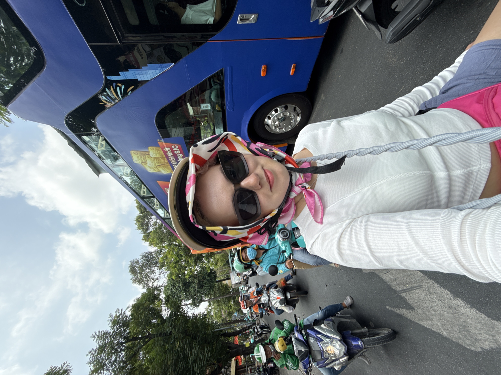 A medium shot depicts a woman in Thủ Đức, Vietnam. She wears a white, long-sleeved shirt, sunglasses, and a colorful headscarf with a hat. She is on the street, surrounded by many motorcycles, and is positioned close to a blue bus. The surroundings include trees and signs.