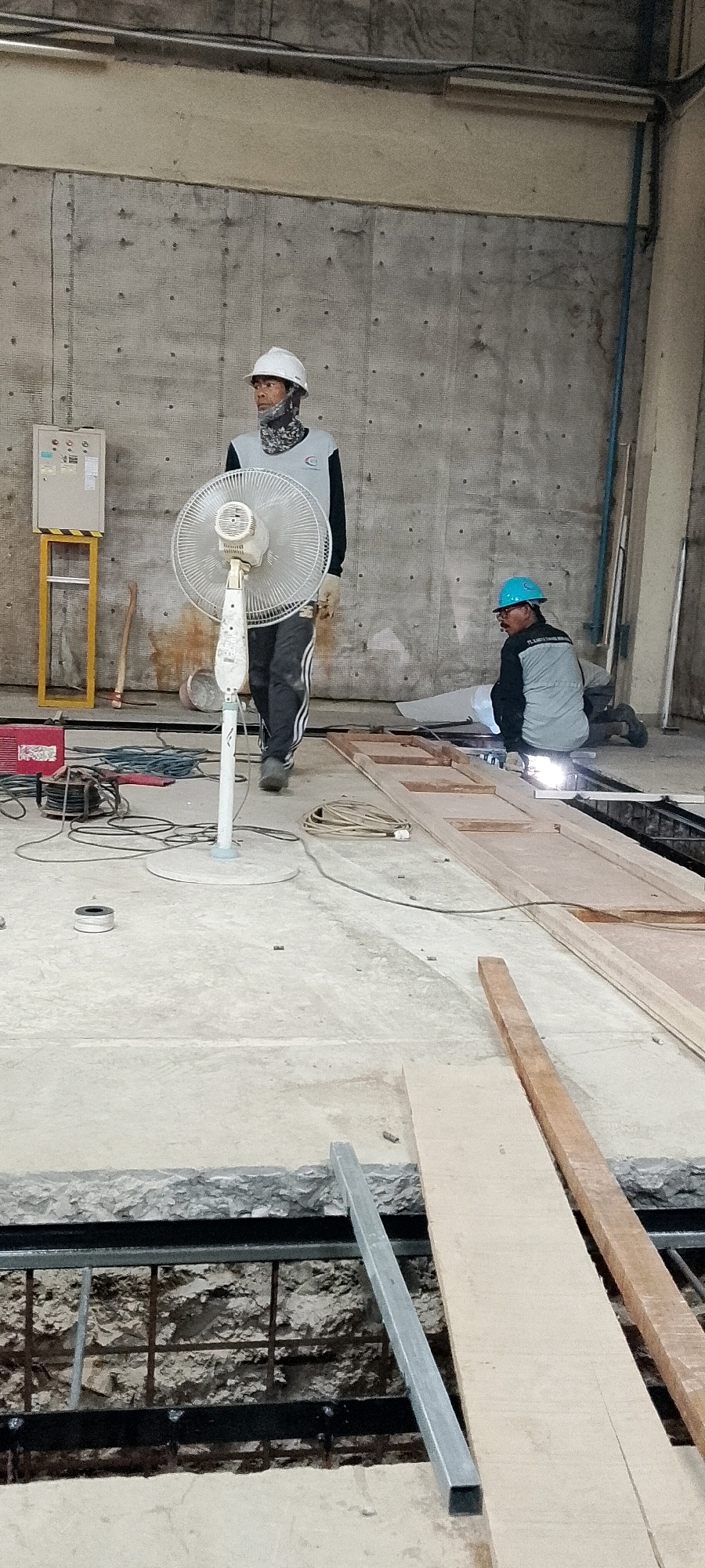 The image depicts an indoor construction site, likely in Depok, Indonesia. Two construction workers are visible. One worker, wearing a white hard hat and a grey and black long-sleeved shirt with black pants, stands near a white oscillating fan. This worker is facing left, looking slightly upwards. The second worker, wearing a blue hard hat and a grey vest over a dark long-sleeved shirt and dark pants, is kneeling on the floor on the right side of the image. This worker is actively welding, indicated by the bright spark emanating from the welding area. The environment is characterized by a concrete floor and a grey concrete wall in the background. Several construction materials, including wooden planks and spools of wire, are scattered around the site. Electrical equipment, including a red welding machine and various cords and cables, are also present. The overall scene suggests ongoing construction or renovation work.