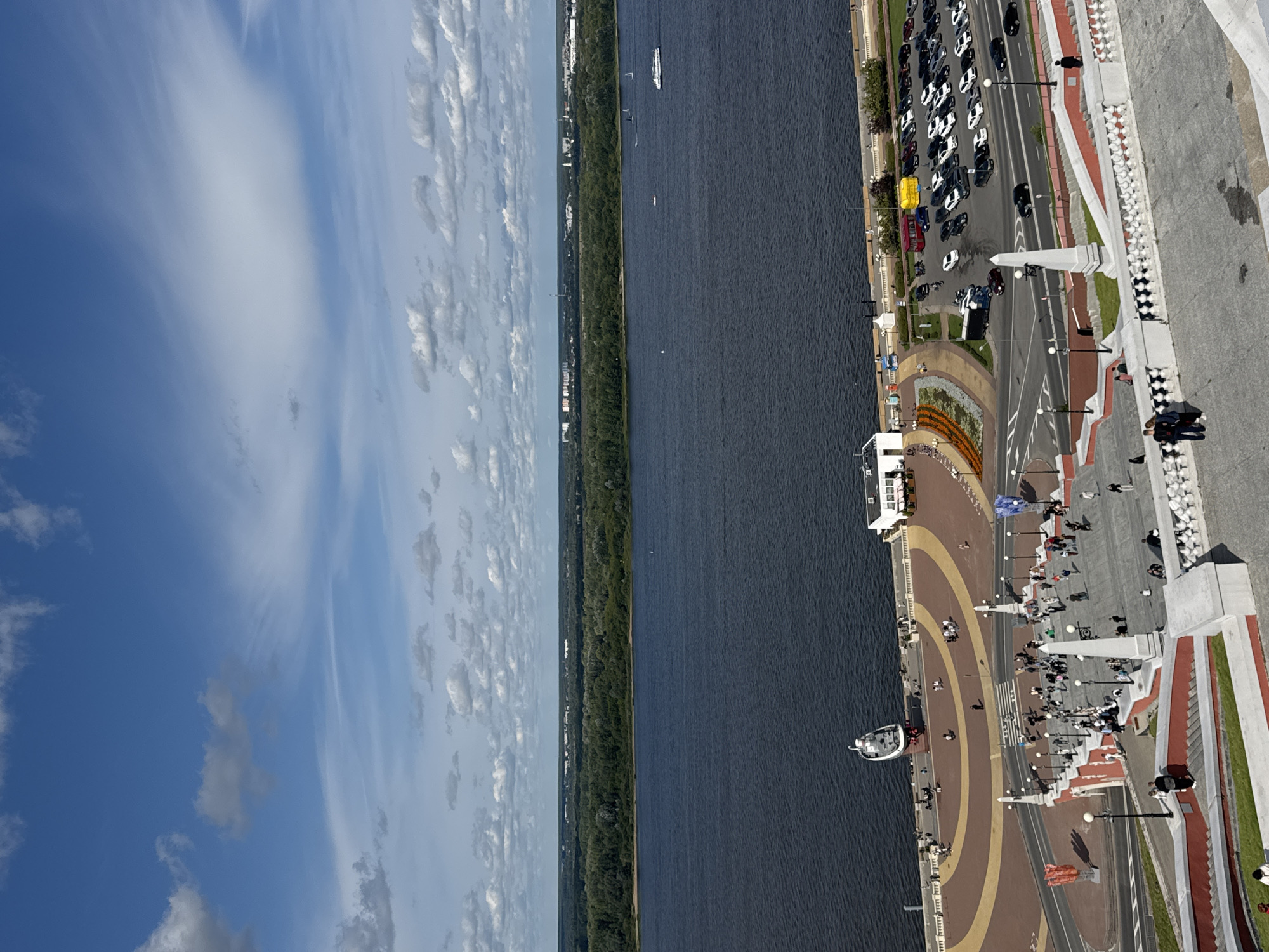 This is a high-angle, full-shot, outdoor, daytime photograph taken in Nizhny Novgorod, Russia. The image captures a view of the confluence of the Volga and Oka rivers, with a wide expanse of dark blue water dominating the middle ground. On the far bank of the river, a dense, green forest stretches across the horizon, dotted with distant buildings and structures.
In the foreground, a large, tiered staircase with white balustrades and red accents descends into the frame. Numerous people are visible on the stairs, some walking, others standing in groups. The stairs lead to a wide, paved embankment area bordering the river.
Along the embankment, a white building with a red roof stands near the water. A large, decorative spiral walkway, paved in red and yellow, is also visible on the embankment. A boat-shaped monument is positioned on this walkway, with people gathered around it. A parking lot filled with various cars, including sedans, SUVs, and a few trucks, is located further down the embankment. A small boat can be seen on the river, as well as a larger white passenger boat moving away from the shore. The sky above is a vibrant blue, with scattered white and grey clouds. The overall impression is a bustling urban waterfront scene on a sunny day.