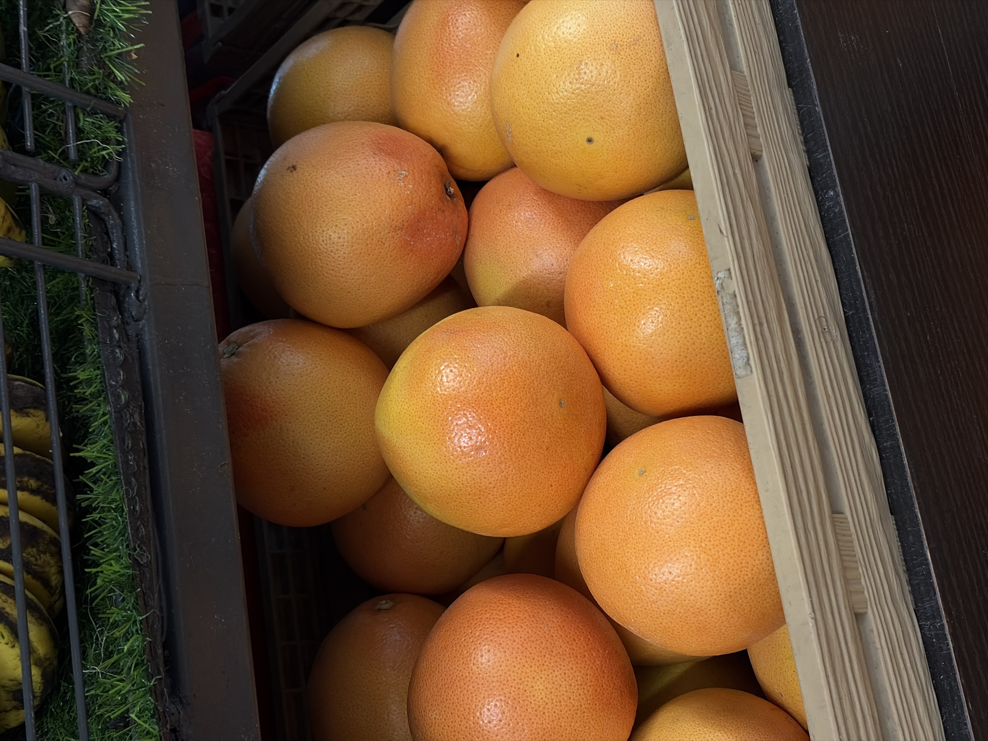 A close-up, eye-level shot shows a wooden crate overflowing with grapefruits. The grapefruits are various shades of orange and yellow, with some showing slight reddish blush. They have a textured peel and some have small brown blemishes. The crate is light brown and appears to be made of weathered wood. In the background, out of focus, are more grapefruits and what looks like a rack of bananas. The lighting is bright, suggesting an indoor setting like a market or grocery store. The scene is devoid of people. The location in Igboefon, Nigeria, is not discernible from the image content.