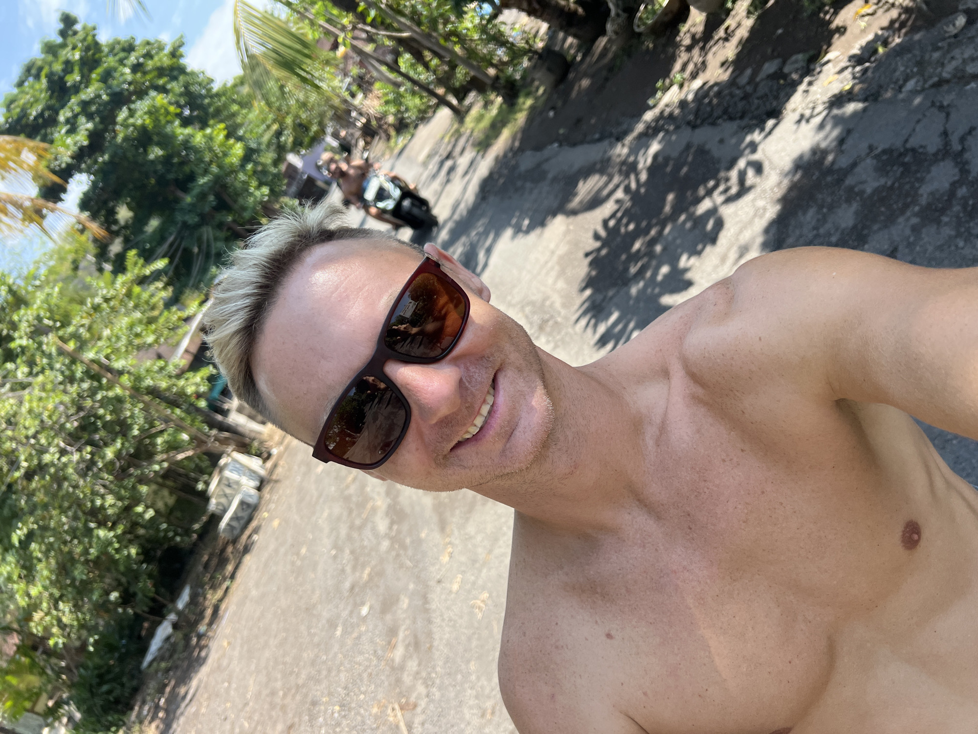 A shirtless man with blond hair and sunglasses smiles at the camera, taking a selfie. He is standing on a paved road lined with trees and lush vegetation. In the background, another person rides a scooter down the road. The scene appears to be in a rural or suburban area of Abang, Indonesia, with sunlight casting shadows on the ground.