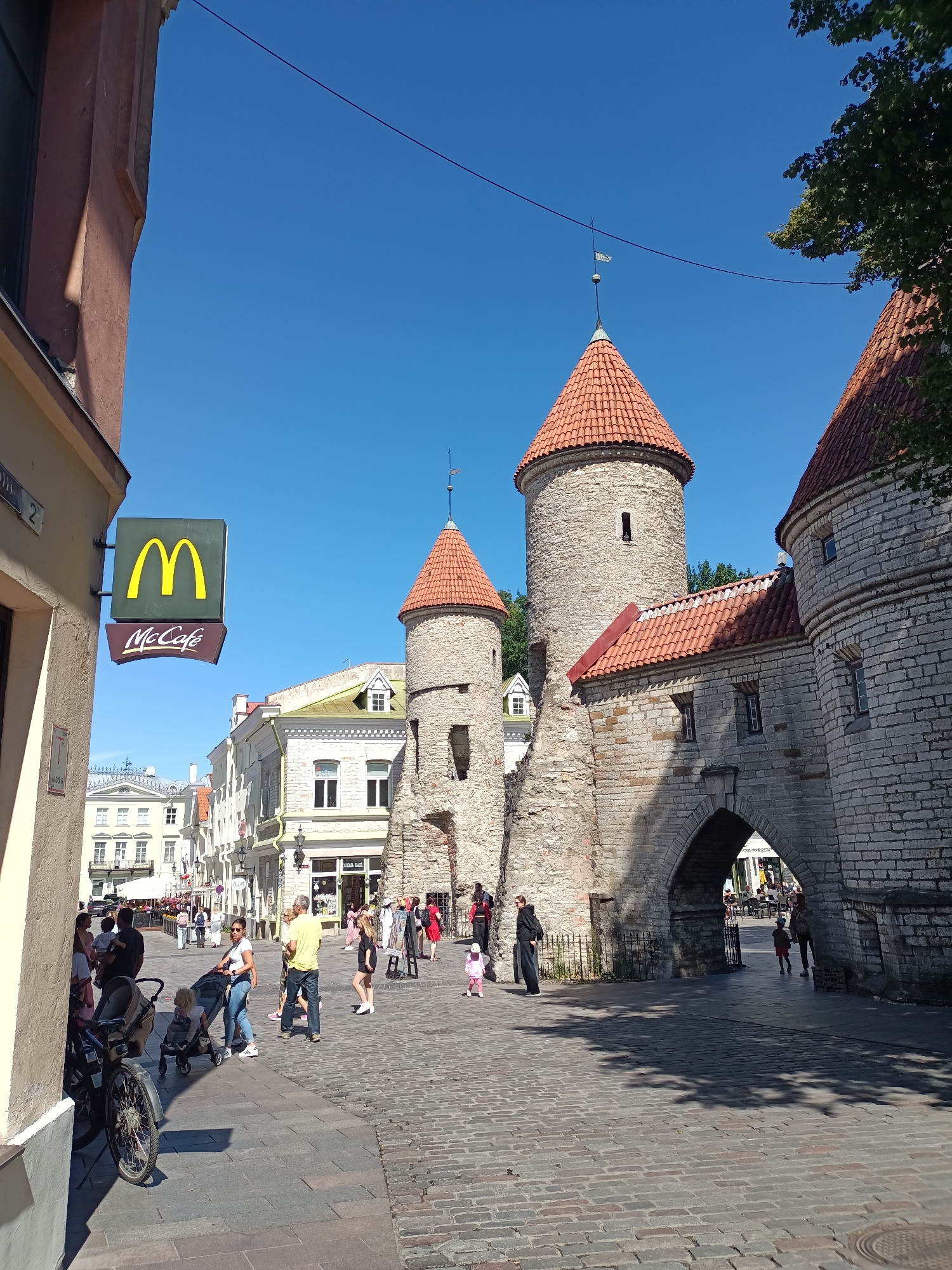 This is a street-level, eye-level, daytime shot of a cobblestone plaza in Tallinn, Estonia, featuring a segment of the historic city wall and several people engaged in various activities.
In the foreground, a section of a tan-colored building with architectural details is visible on the left. A prominent McDonald's sign, featuring the golden arches and the "McCafé" logo, is mounted on this building. To the right of the McDonald's sign, the ancient stone city wall of Tallinn is present, characterized by its rough-hewn grey stone blocks and reddish-tiled conical turrets. An arched gateway is visible within the wall, allowing passage to a street lined with buildings beyond.
Several individuals are scattered across the plaza. On the left side of the plaza, near the McDonald's, a group of people are gathered. A woman is pushing a stroller, and a bicycle is parked nearby. Further into the plaza, more people are walking and conversing, including a woman with a child in a stroller and a man in a yellow shirt.
In the mid-ground, the stone turrets of the city wall rise majestically, with their tiled roofs contrasting against the clear blue sky. The architecture of the buildings lining the street behind the wall appears to be a mix of historical styles, with light-colored facades and red-tiled roofs.
The overall scene depicts a bustling public space where historical architecture meets modern life. The lighting suggests a bright, sunny day.