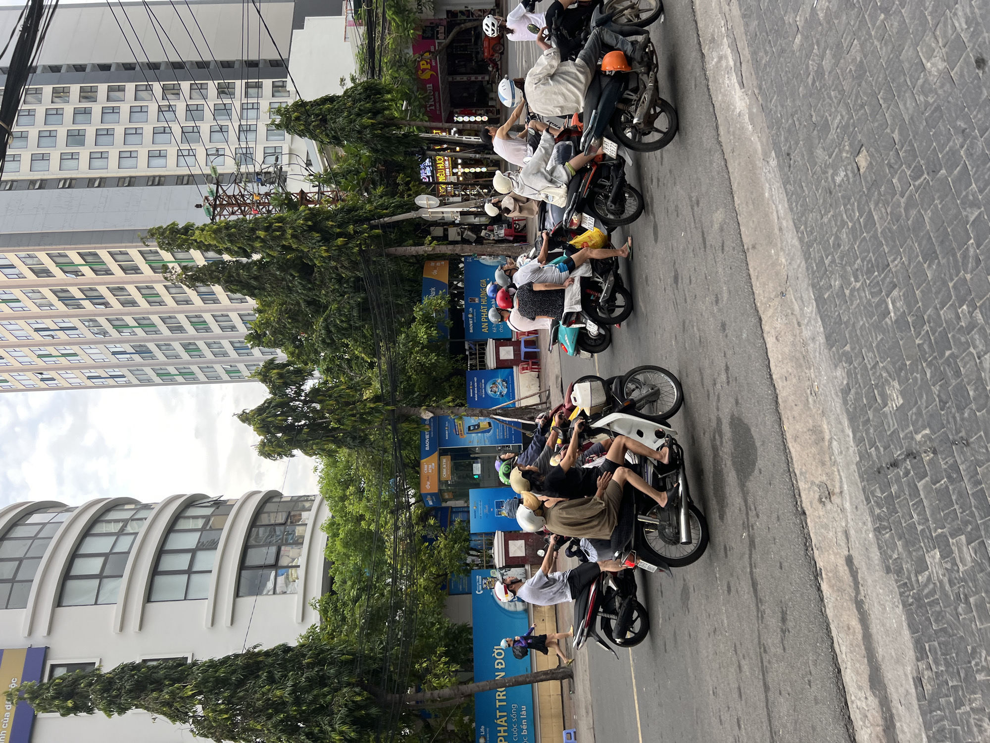 The image depicts a street scene in Nha Trang, Vietnam, characterized by a traffic jam of motorcycles. In the foreground, several individuals are riding motorcycles, navigating the crowded street. The vehicles are varied in color and design, with some carrying multiple passengers. The people on the motorcycles are mostly wearing helmets, suggesting adherence to local traffic laws. The background features tall buildings, typical of an urban environment, along with trees lining the street. The road surface appears to be asphalt, with some sections showing signs of wear. The overall atmosphere suggests a busy, bustling city environment common in Vietnam.