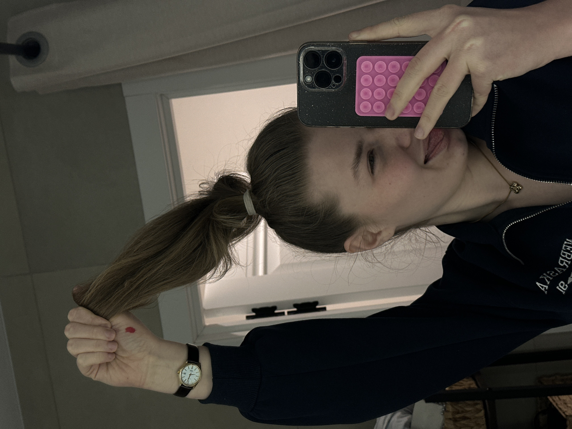 A person is taking a selfie in what appears to be a bathroom. The person is holding a phone with a pink pop socket and sticking their tongue out. The person's hair is pulled back in a ponytail. They are wearing a dark blue zippered jacket and a watch with a black band. There is a small red mark on their hand. The background includes a mirror, part of a window, a curtain rod, and light-colored walls. The location cannot be determined from this image.