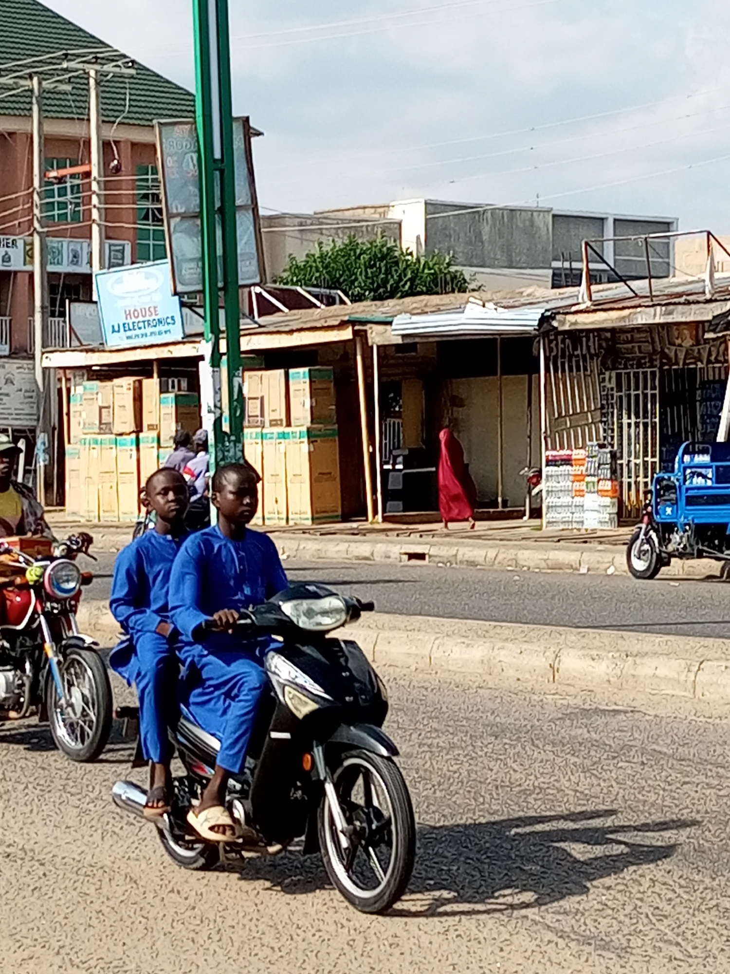 In Arawa, Nigeria, on a sunlit day, two young boys in matching blue traditional attire ride a black motorbike down a paved road. They are positioned side-by-side, with one boy in front operating the handlebars and the other seated behind him. Their feet, clad in light-colored sandals, are visible resting on the footpegs. The motorbike is in motion, casting distinct shadows on the textured asphalt.
To the left, a red motorbike is parked, its front wheel visible. In the background, a street scene unfolds with several buildings and shops. A prominent green pole stands on the sidewalk. A blue sign advertising "HOUSE AJ ELECTRONICS" is mounted on one of the buildings. Stacks of large cardboard boxes are visible outside some of the shops. Further down the street, a small blue truck with a cargo bed is parked. A person in a flowing red garment walks on the sidewalk. The sky above is overcast with faint clouds.