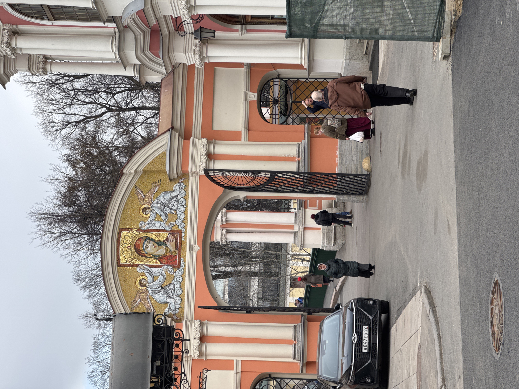 The image depicts a street scene in Saint Petersburg, Russia, with a decorative archway as a focal point. The archway is constructed with red and white columns and features a mosaic of a religious icon on top. Several people are present: a couple near the archway's upper portion, one person walking through the arch, and another figure further down the street. A black car is parked near the archway, visible on the right side of the image. Bare tree branches are visible above the archway. The weather appears overcast.