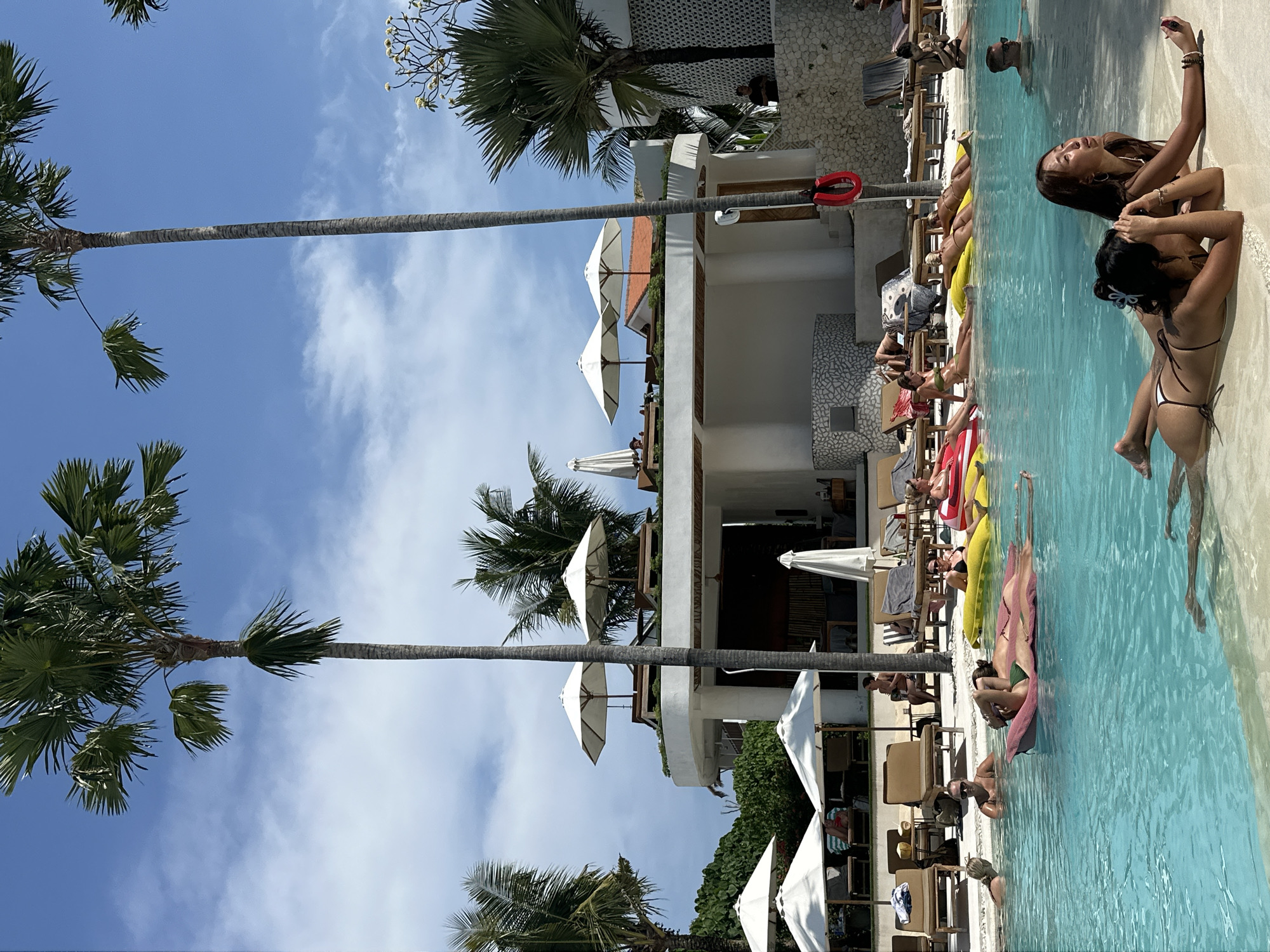 The image depicts a bustling poolside scene in North Kuta, Indonesia. The composition showcases a variety of individuals engaging in leisurely activities.
In the foreground, several women are lounging in the pool, some with their bodies partially submerged. One woman is taking a photo.
The pool is bordered by lounge chairs, many of which are occupied by sunbathers. Umbrellas are strategically placed to offer shade to the patrons.
In the background, a two-story building adds architectural interest. Tall palm trees are interspersed throughout the scene. The sky is bright blue, with scattered clouds.