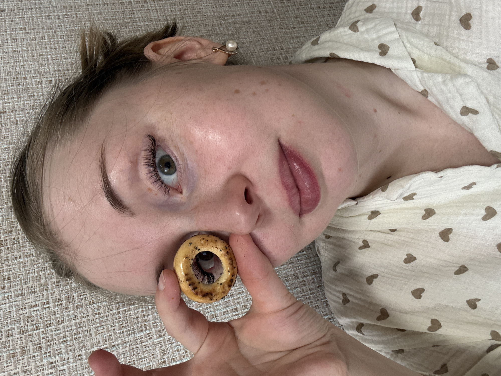 A woman is lying on a tan fabric surface, holding a small bagel over her eye. She is wearing a light-colored top with brown hearts and a pearl earring.