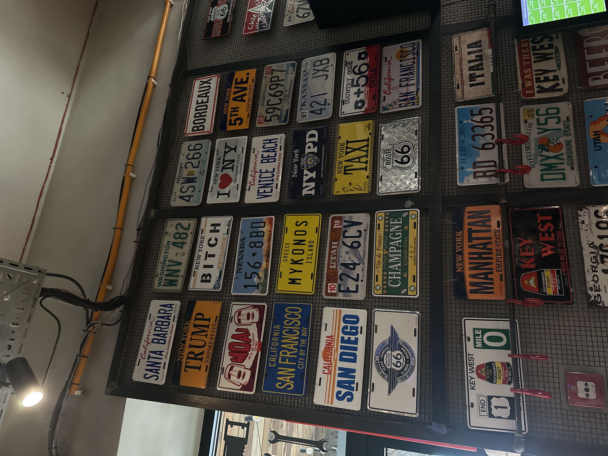 A wall is decorated with a collection of novelty license plates. The plates originate from various states and countries and are attached to a metal grid. The plates feature text, state names, and slogans. The license plates are mounted on the wall of an indoor space, which is dimly lit by a spotlight. Some plates are mounted with red, pick-like clips. The location in the city of Krasnoyarsk, Russia is not apparent from this image.