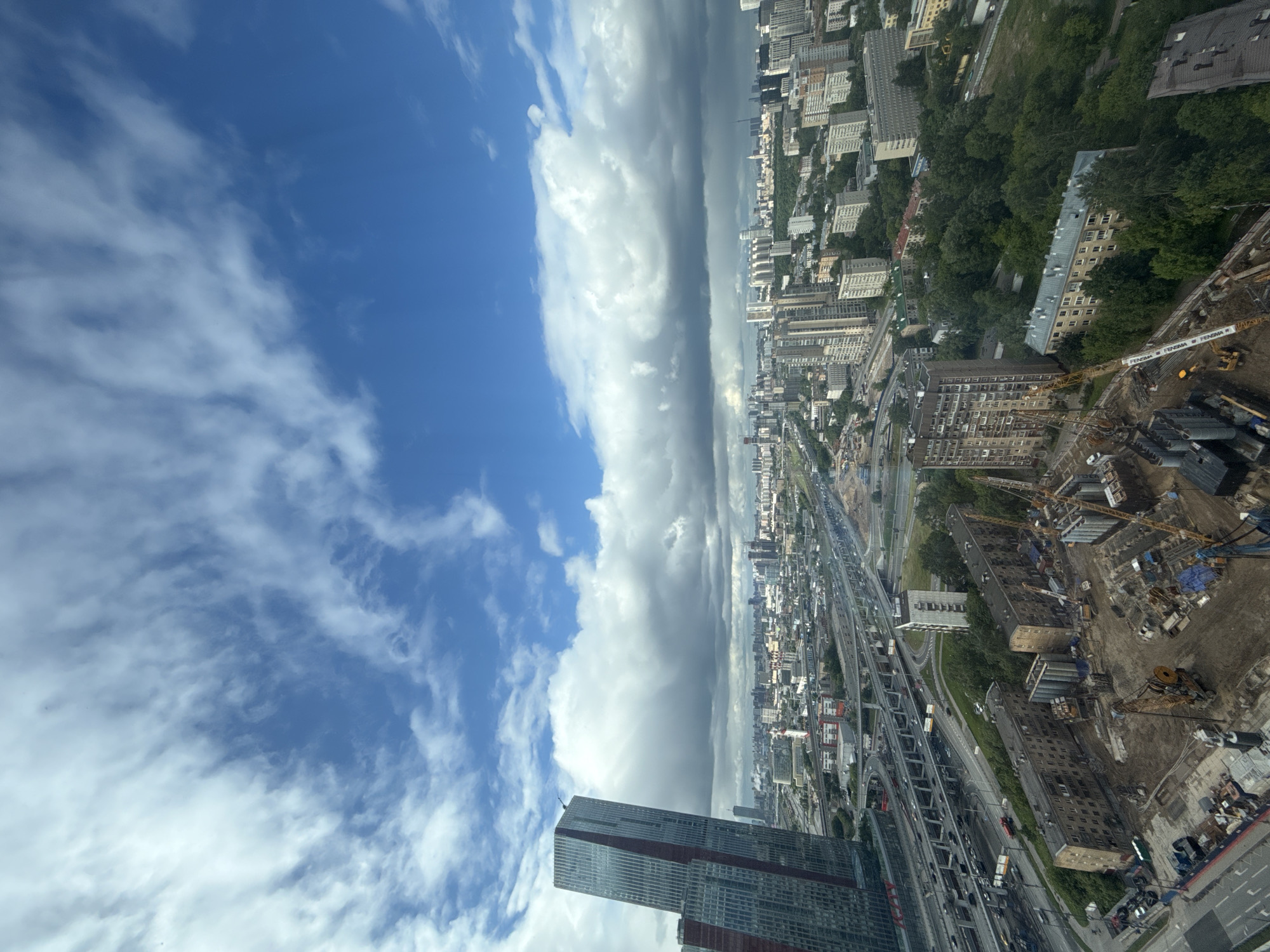 The image presents a high-angle view of what appears to be a cityscape in Khimki, Russia, observed from a significant altitude, possibly a high-rise building or an aircraft. The sky dominates a substantial portion of the frame, featuring a contrast of clear blue interspersed with patchy, white clouds on the left, shifting to a dense, gray cloud formation in the central area.
Below, the urban landscape unfolds. A network of roads and highways is visible, with multiple overpasses and a moderate density of vehicular traffic. A mix of residential and commercial buildings is discernible, ranging in height and architectural style. A large construction site is present, characterized by construction equipment, unfinished structures, and building materials.
In the lower portion of the picture, parts of a tall, modern skyscraper with a glass façade and red accents can be seen.