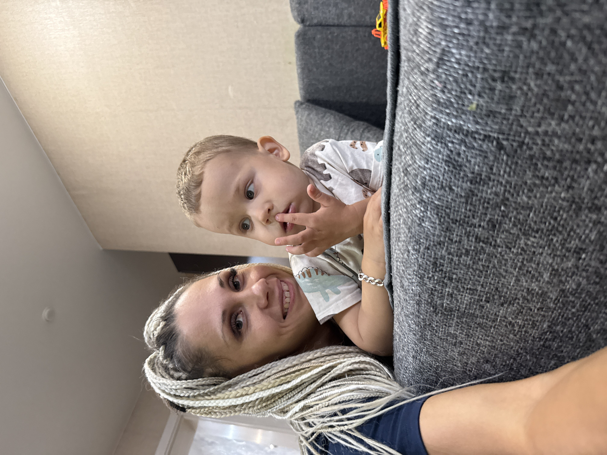 A medium shot captures a woman with blonde braids and a young boy on a gray sofa. The woman is on the left, looking towards the camera with a smile. Her hair is braided and falls over her shoulder. The boy is to her right, looking away from the camera with his finger near his mouth. He is wearing a white t-shirt with a print. A small yellow toy car is visible on the sofa to the right of the boy. The background consists of a textured wall and a ceiling. The image appears to be taken indoors, likely in a residential setting in Rusanovo, Russia, as indicated by the provided context.