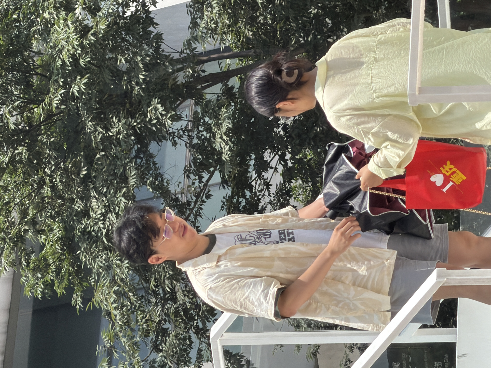 In Yuzhong District, China, on a bright, sunny day with a lush green tree providing shade, a young man and woman stand on a white staircase. The man, wearing a cream-colored patterned shirt over a white graphic tee and gray shorts, is looking down and holding a black umbrella. To his right, a woman in a pale yellow shirt with her hair pulled back in a bun is holding a bright red tote bag with gold chain straps. The bag features Chinese characters and a white heart emblem. The background shows a modern building and dense foliage.