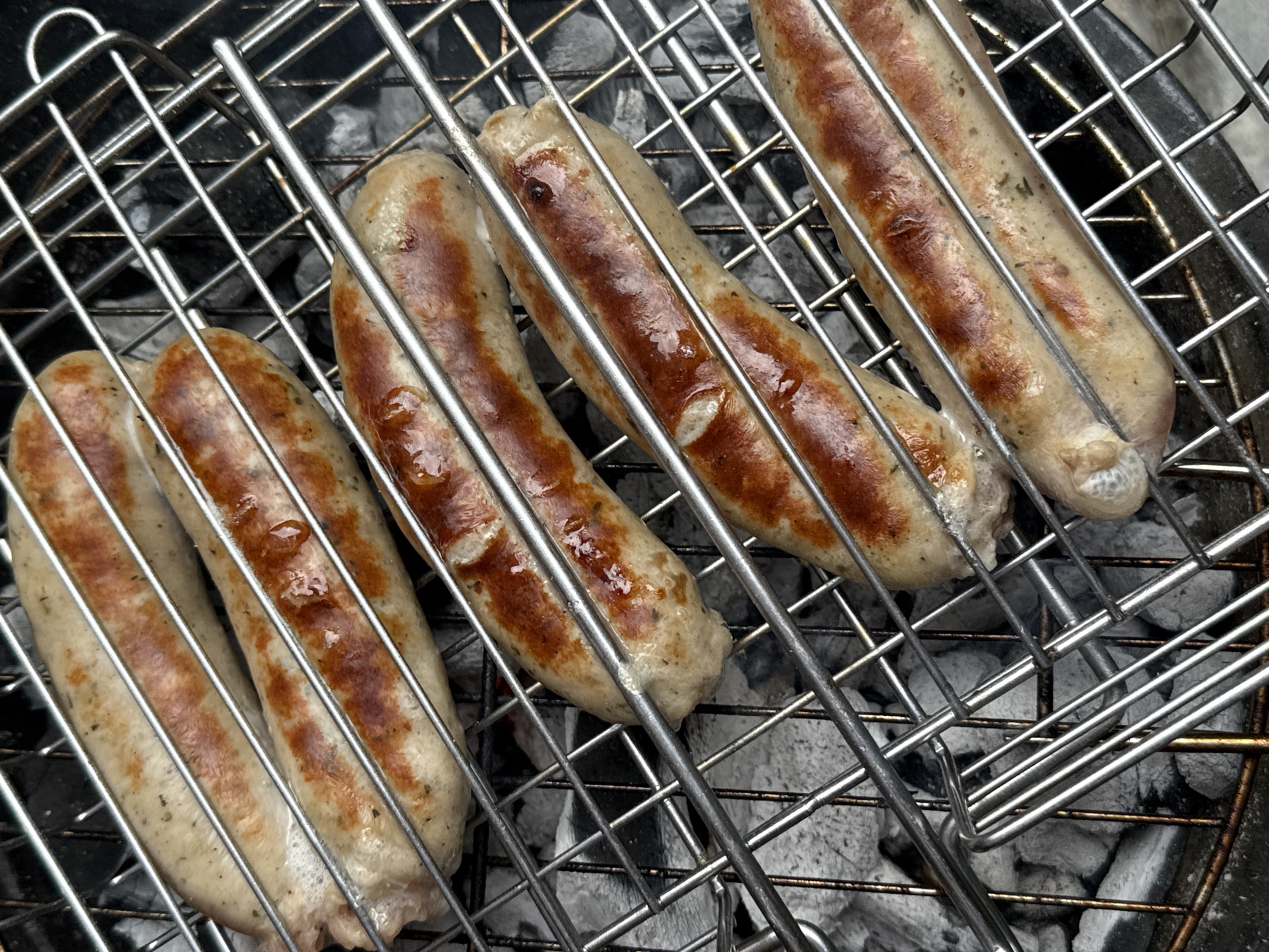 The image shows four sausages grilling on a metal rack over hot coals. The sausages are plump and have a golden-brown, slightly charred exterior. They are arranged diagonally across the frame, with the top sausage being the closest to the viewer. The grilling rack is made of silver-colored metal bars. The background consists of gray, glowing coals. The scene is shot from a top-down perspective. This image does not contain any information about the city of Wrocław, Poland.