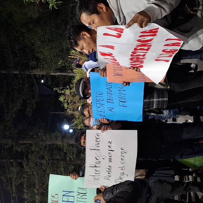 A group of people are protesting at night, holding signs with messages in Spanish. One sign reads "Victor Hugo Rivera, Alcalde Incapaz, Deja el Cargo" (Victor Hugo Rivera, Incapable Mayor, Leave the Post). Another says "Respeto al Trabajo Formal" (Respect for Formal Work). A third sign criticizes "competencia ilegal" (illegal competition). It appears to be a demonstration against a mayor and in support of formal employment.